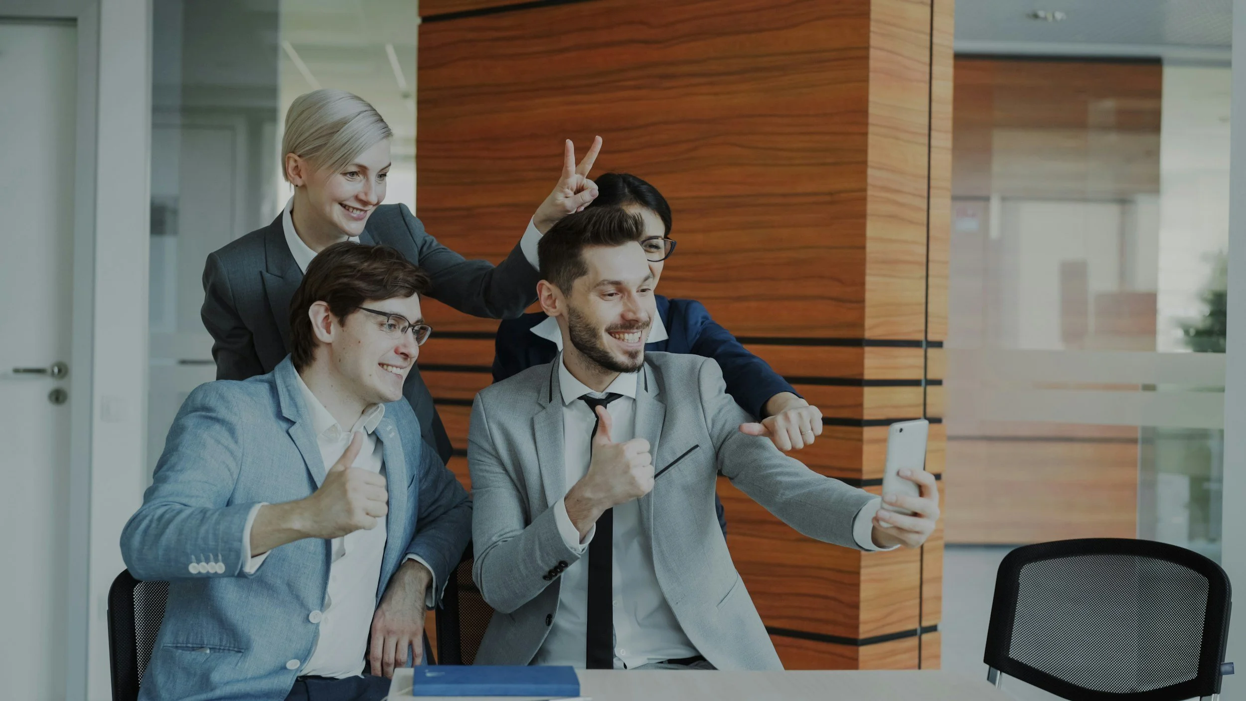 Four business professionals taking a selfie in an office, all smiling and giving thumbs up, with two women and two men, one of the women has short blonde hair and the other has short black hair, one of the men has glasses and dark hair, the other has light hair and a beard.