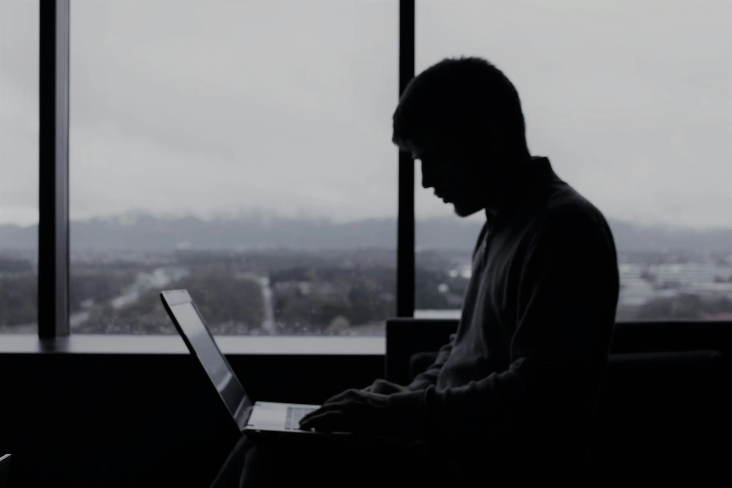 Silhouette of a person working on a laptop in an office with large windows showing a cloudy sky and cityscape in the background.