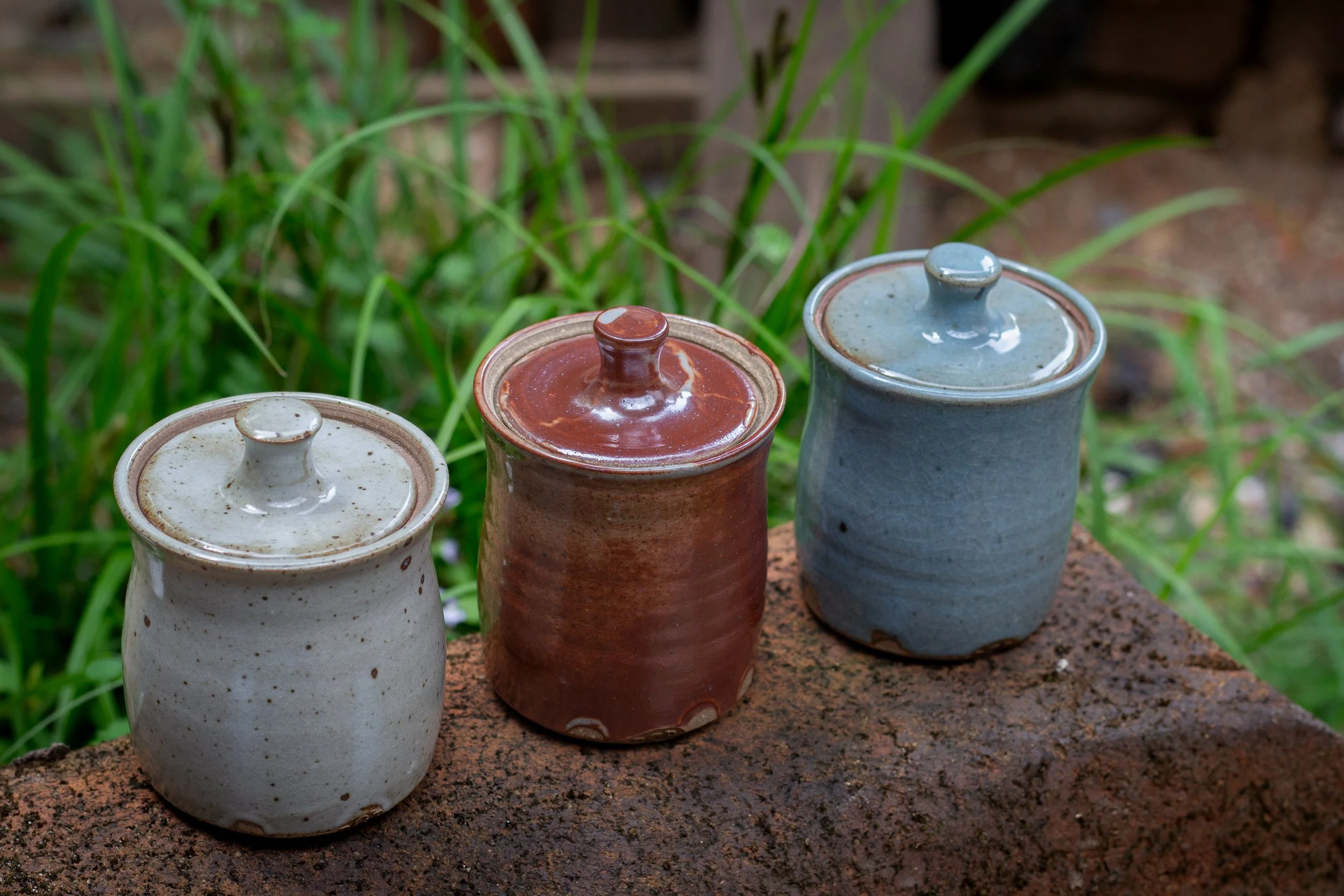 Three ceramic jars with lids, arranged outdoors on a rock surface, with green grass and plants in the background.