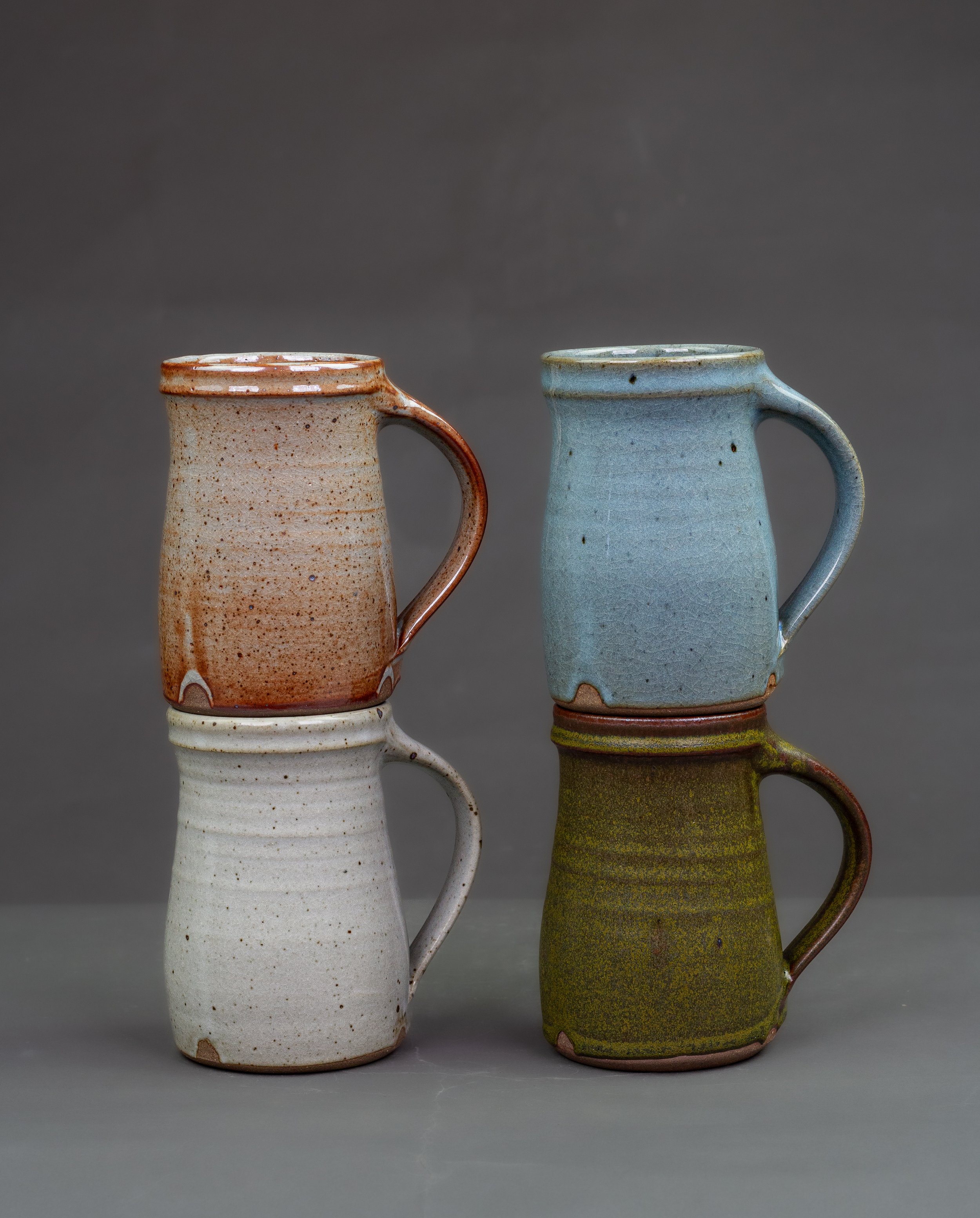 Four ceramic tankards with handles, one in earthy orange-brown glaze and the other in matte green tenmoku glaze, one cream dolomite glaze, on blur Chun glaze both with textured surfaces, placed side by side on a grey background.