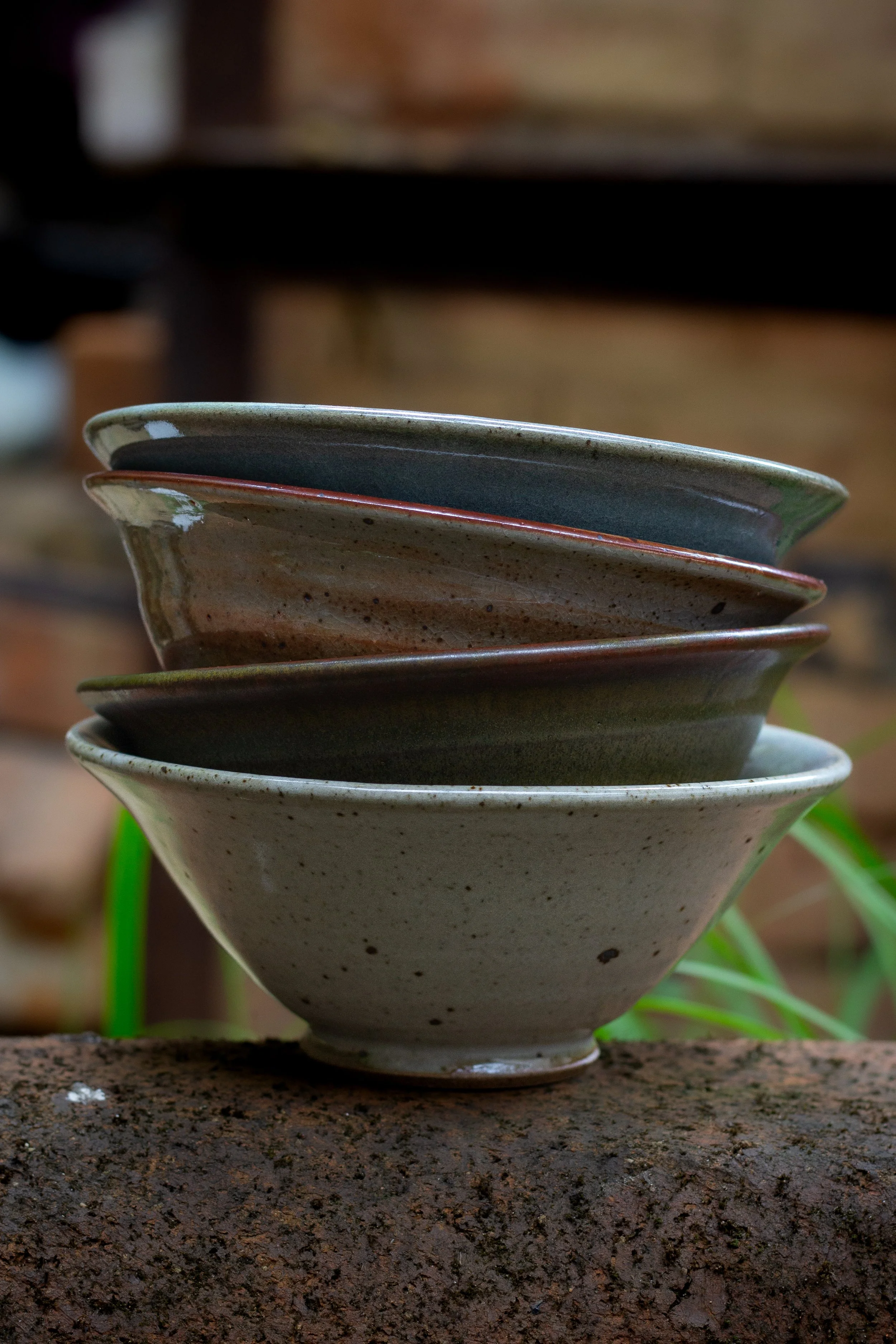 Close-up of a stack of pottery bowls with earthy tones, placed outdoors on a rust-colored surface with a blurred background of green plants and a brick surface.