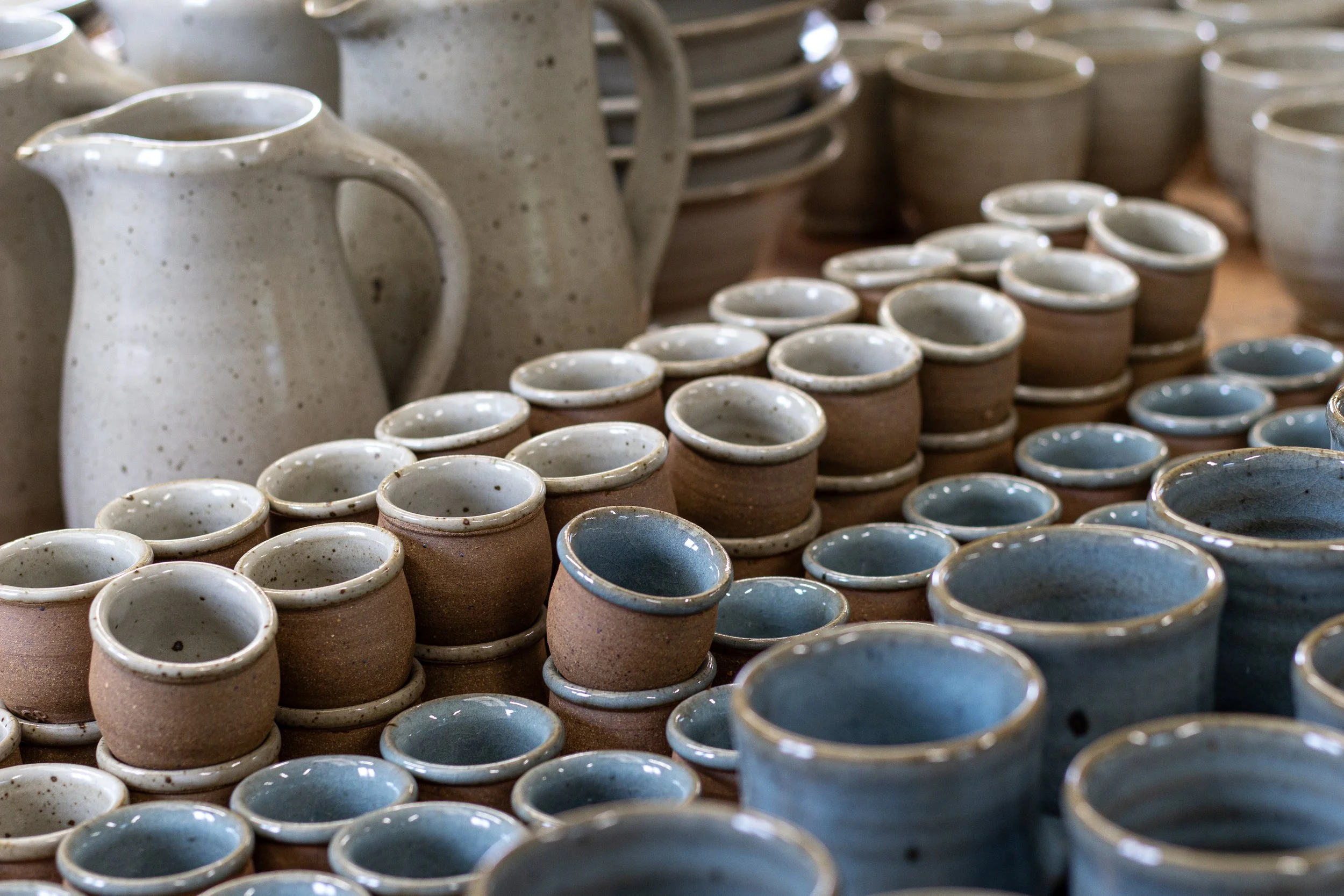 Multiple ceramic pottery pieces, including cups, bowls, and pitchers, displayed in a pottery studio or shop.