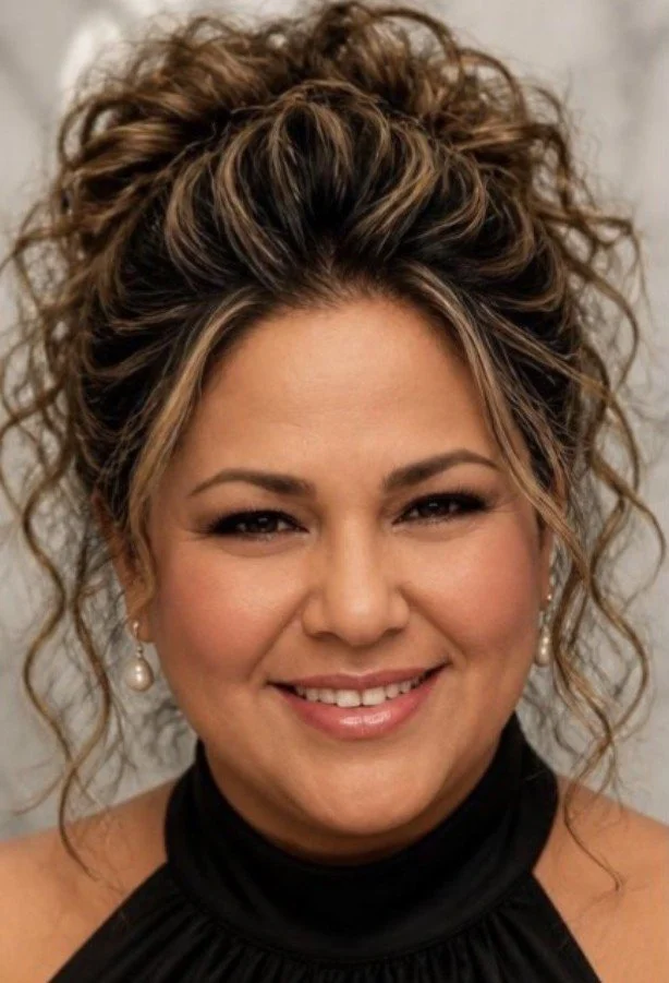 Close-up portrait of a woman with curly brown hair styled in an updo, wearing pearl earrings and a black high-neck top, smiling at the camera.