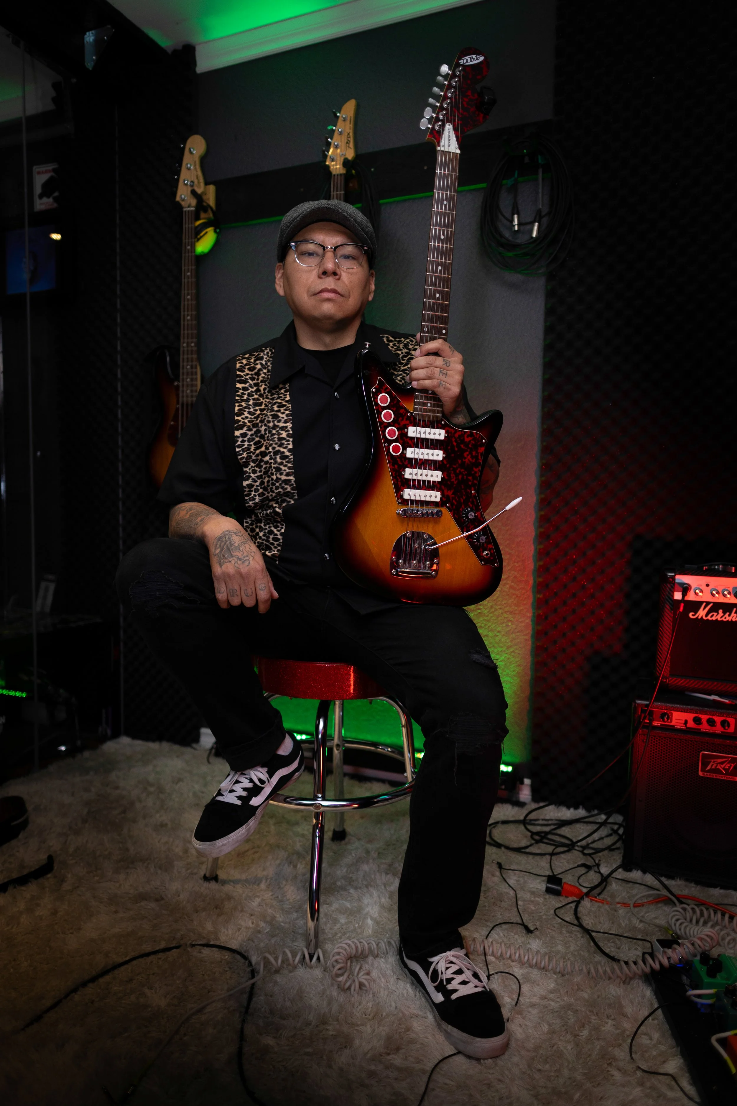 A man sitting on a stool holding an electric guitar in a music studio with musical equipment and hanging guitar on the wall behind him.