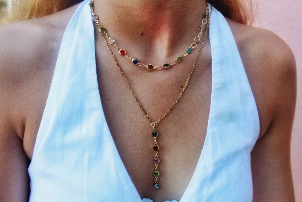 Close-up of a woman wearing three layered gold necklaces with colorful gemstone charms, and a white sleeveless top.