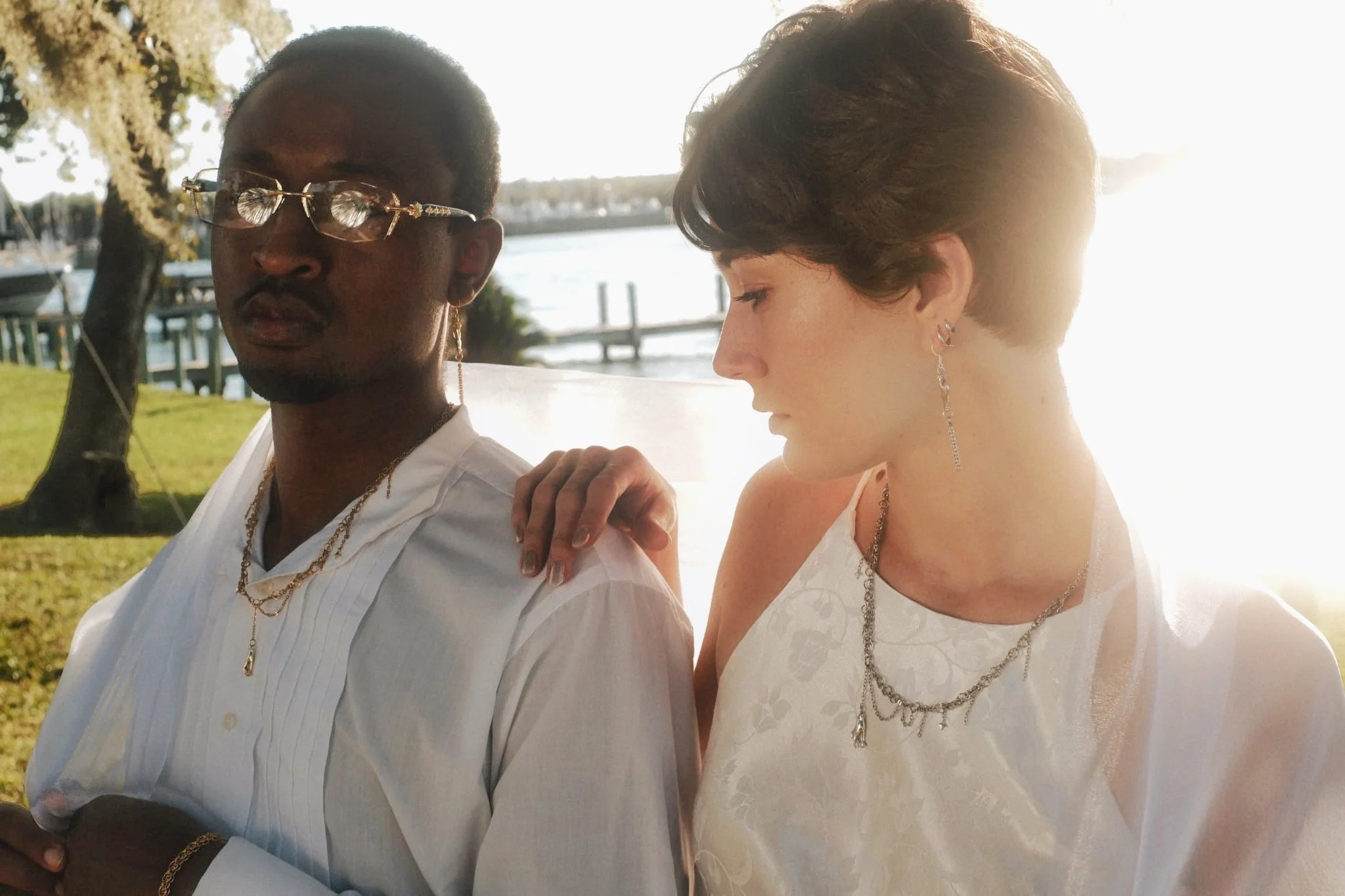 A man and a woman standing outdoors near a body of water, dressed in white, with jewelry, and illuminated by bright sunlight.