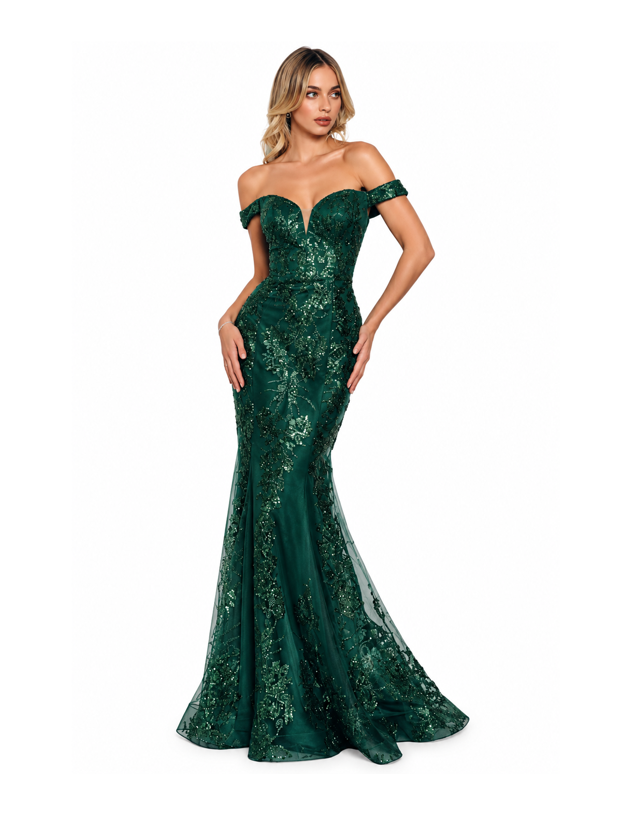 A woman in an emerald green evening gown with off-shoulder design and intricate embroidery posing with hand on hip against a plain white background.