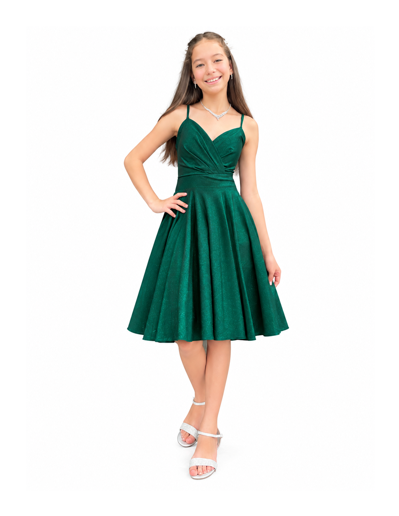 A young woman with long brown hair wearing a dark green, sleeveless, knee-length dress with a fitted bodice and a full skirt, standing with one hand on her hip, smiling, and wearing jewelry including earrings, a necklace, and white high-heeled sandals.