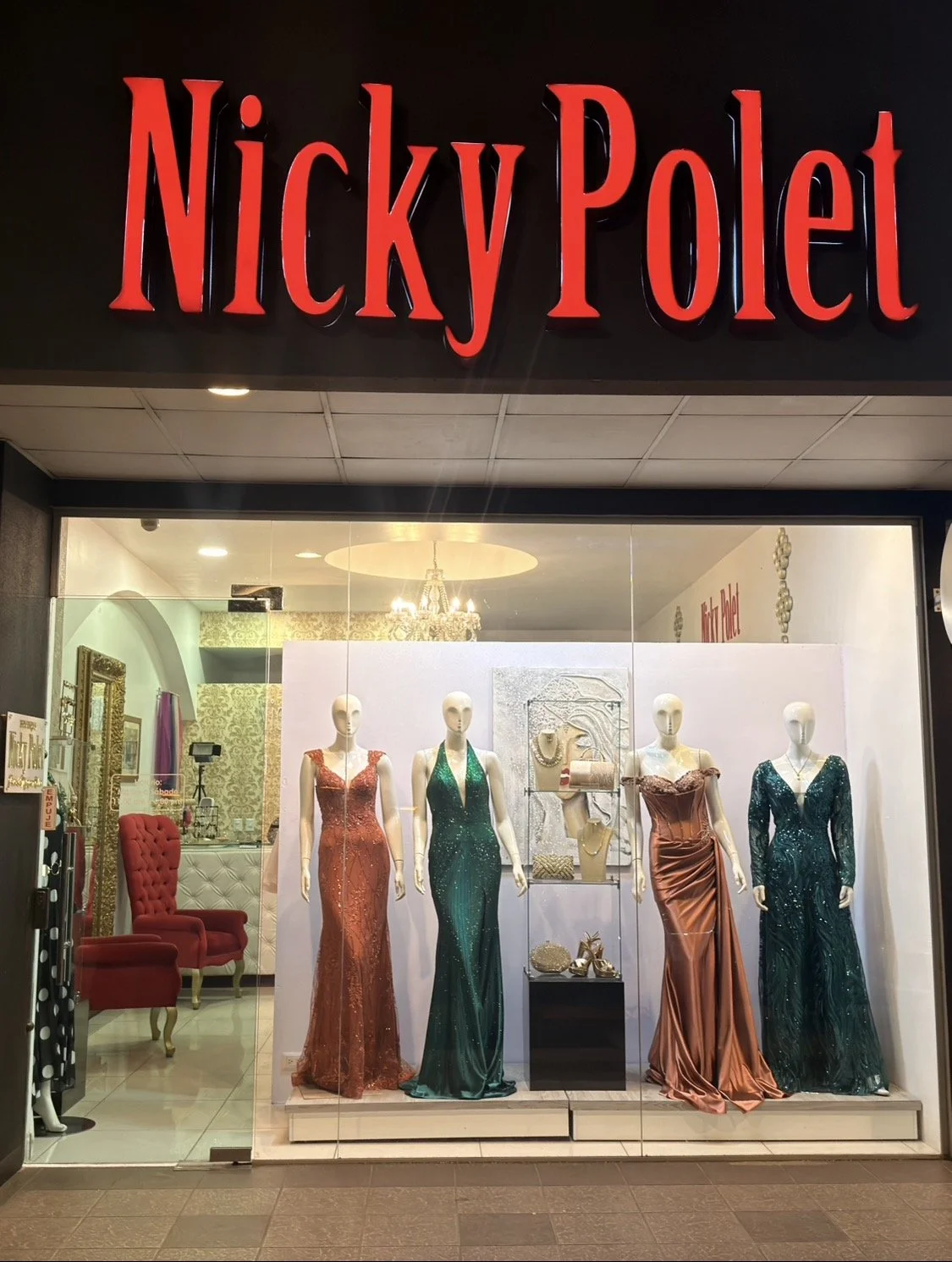 Storefront display window of Nicky Polet with four mannequins dressed in evening gowns. The mannequins are wearing long, elegant dresses in shades of orange, green, and dark teal, with jewelry and accessories. The shop's interior is decorated with a chandelier, a gold-framed mirror, and ornate wallpaper.