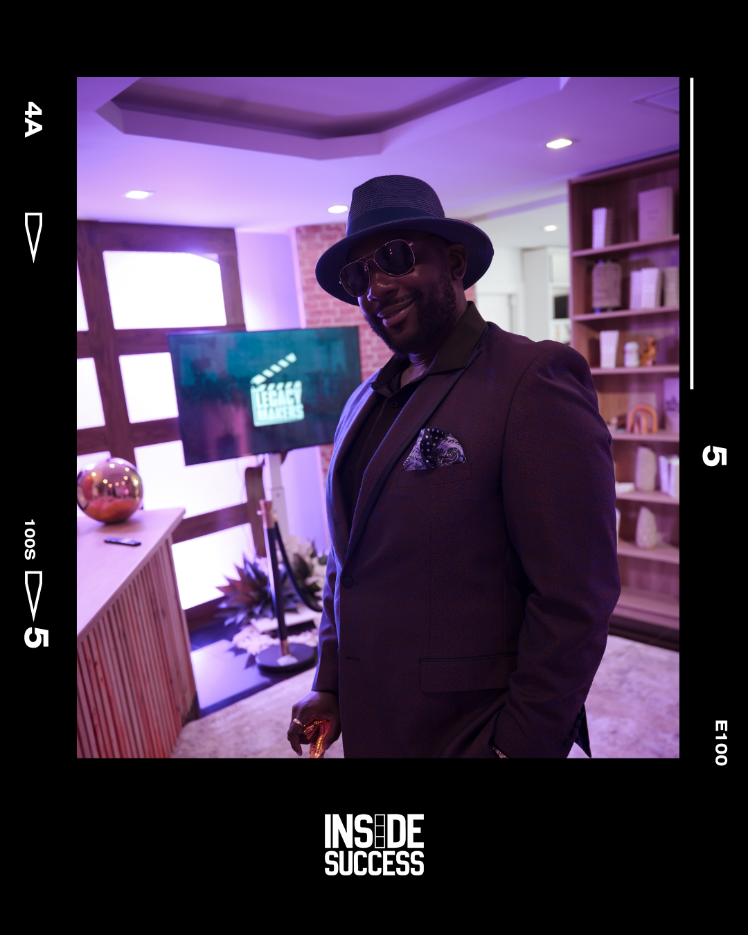 A man in a suit, sunglasses, and a fedora hat smiling indoors with purple lighting, a bookshelf, and a television in the background.