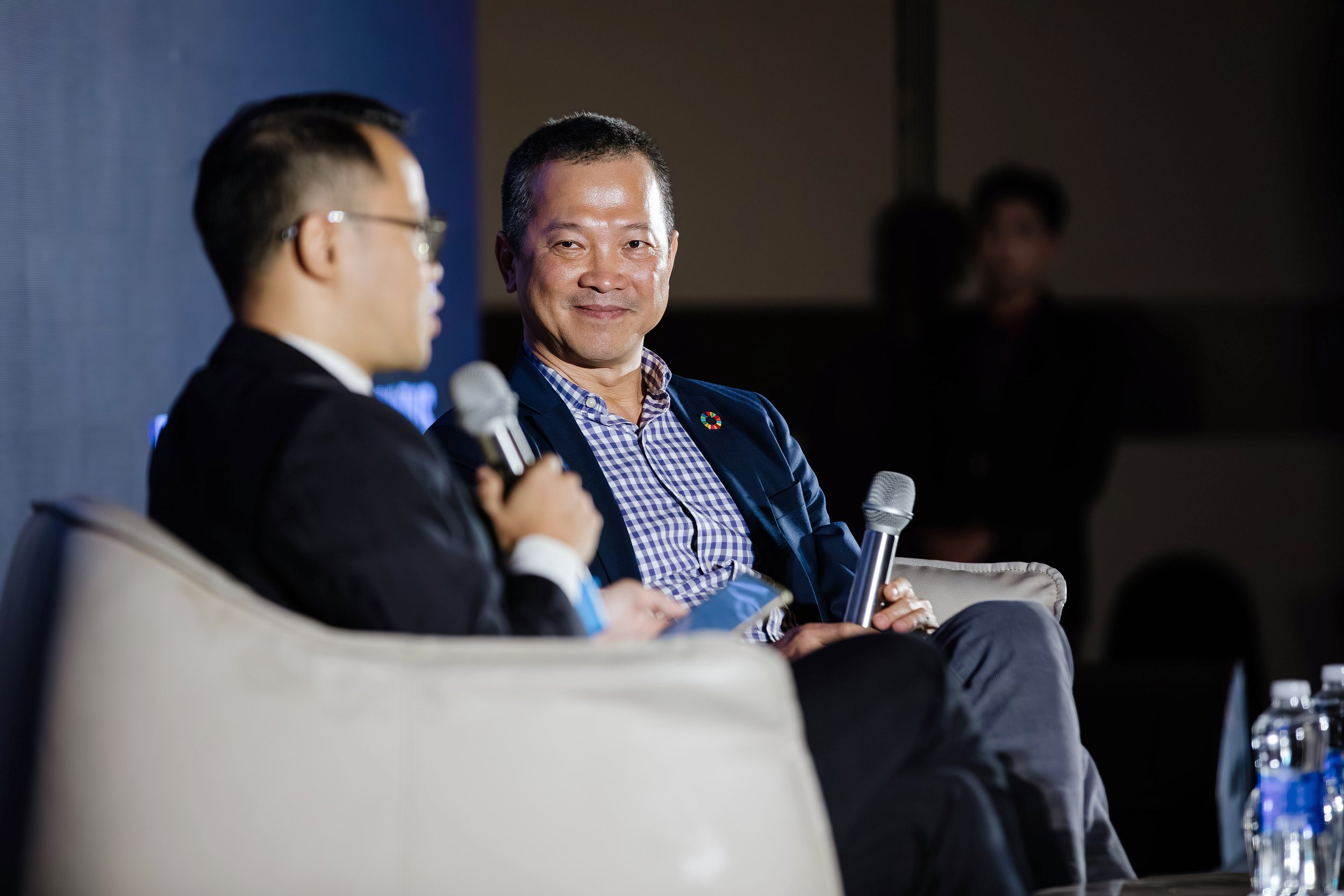 Two men sitting on a stage, holding microphones, engaged in conversation. The man on the right is smiling, wearing a checkered shirt and blazer; the man on the left is talking, wearing a black suit and glasses.