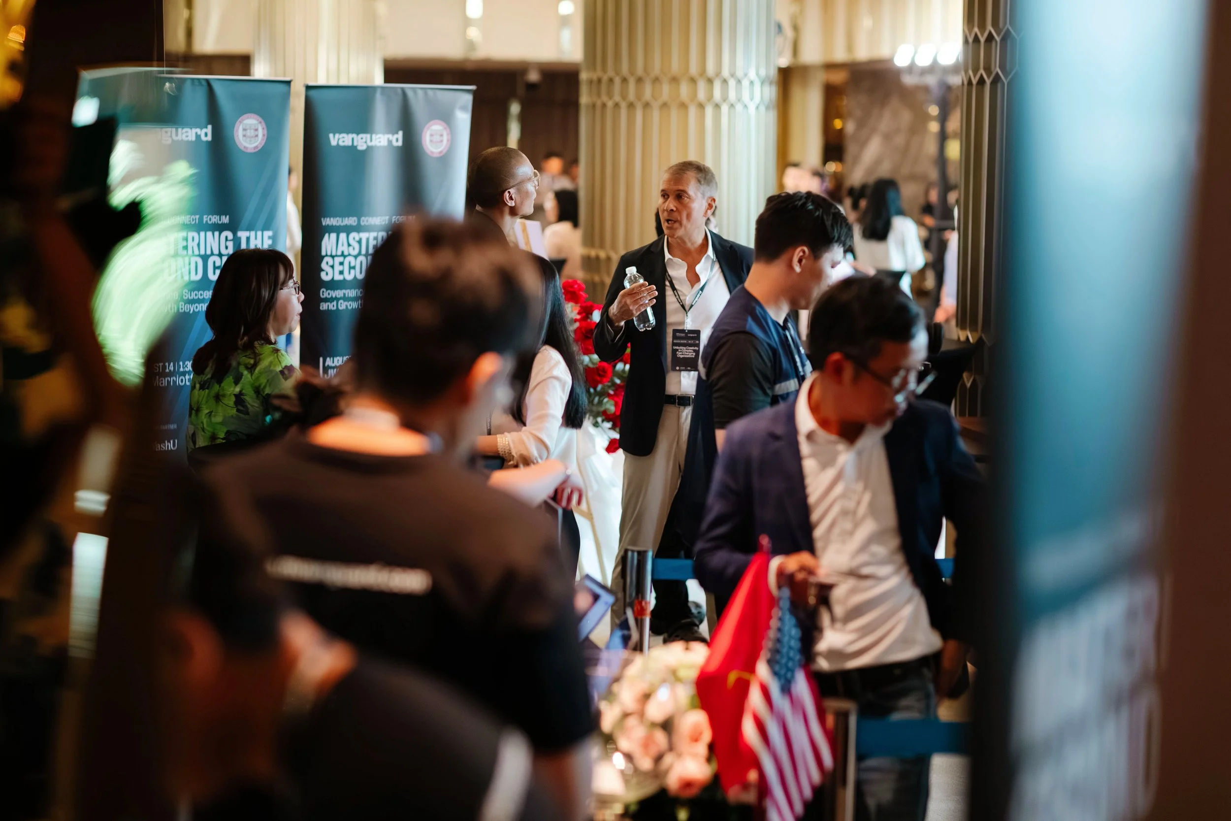 A busy conference with attendees, some talking, some looking at their phones, and national flags displayed among the crowd, with banners stating 'Vanguard' and 'Mastering the Second,' in an indoor venue.