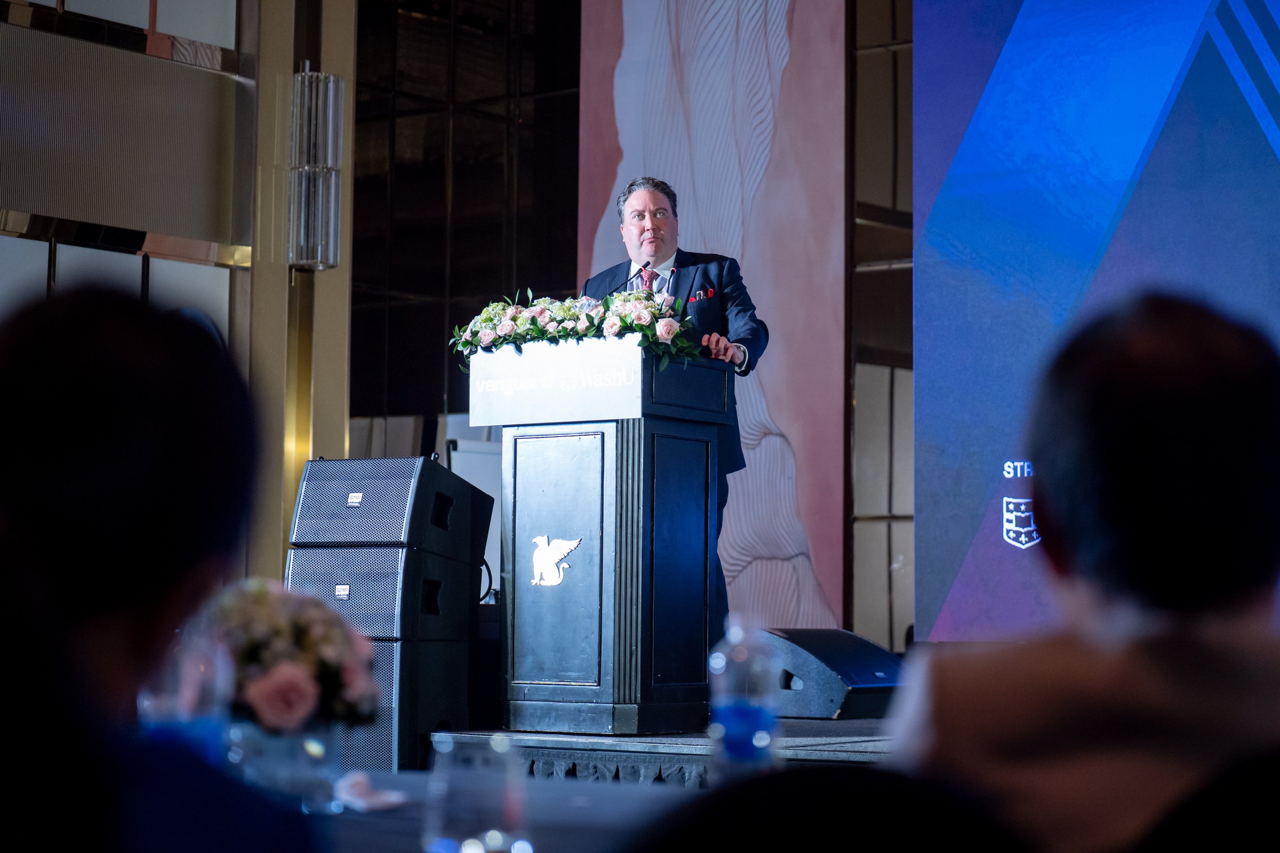 A man in a suit standing behind a podium decorated with pink and white flowers at a formal event, with a large screen and audience in the background.