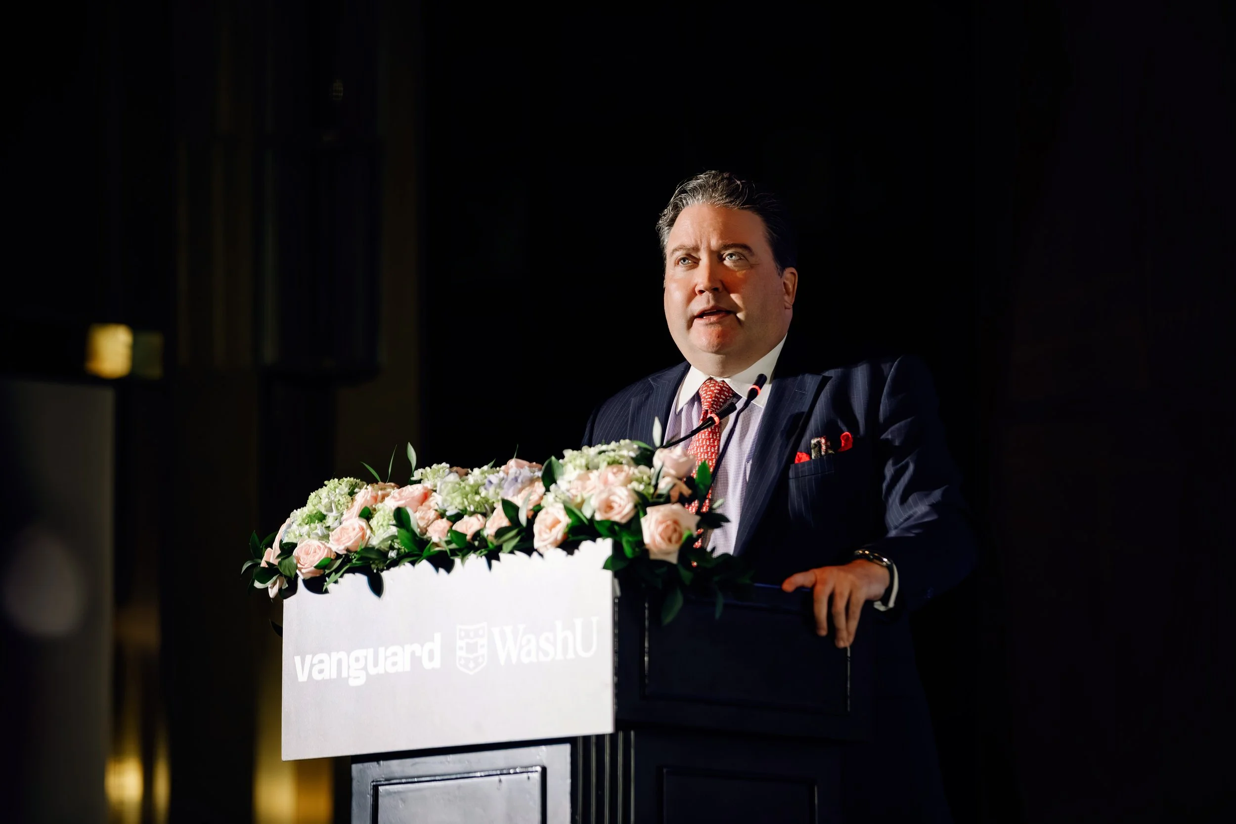 A man in a dark suit with a striped shirt and red tie is standing at a podium decorated with pink and white flowers, delivering a speech. The podium displays the logos of Vanguard and WashU.