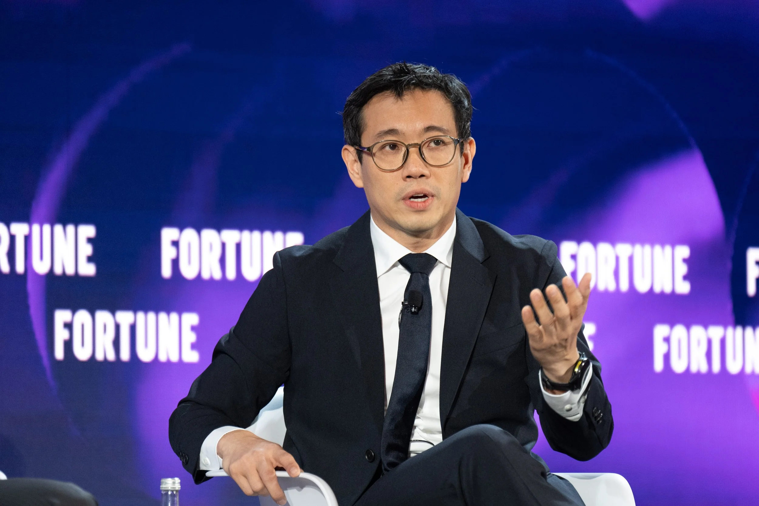 A man in a black suit and glasses speaking into a microphone at a conference or event. Behind him is a partially visible large blue screen with the words 'MAC SEC' and additional text.