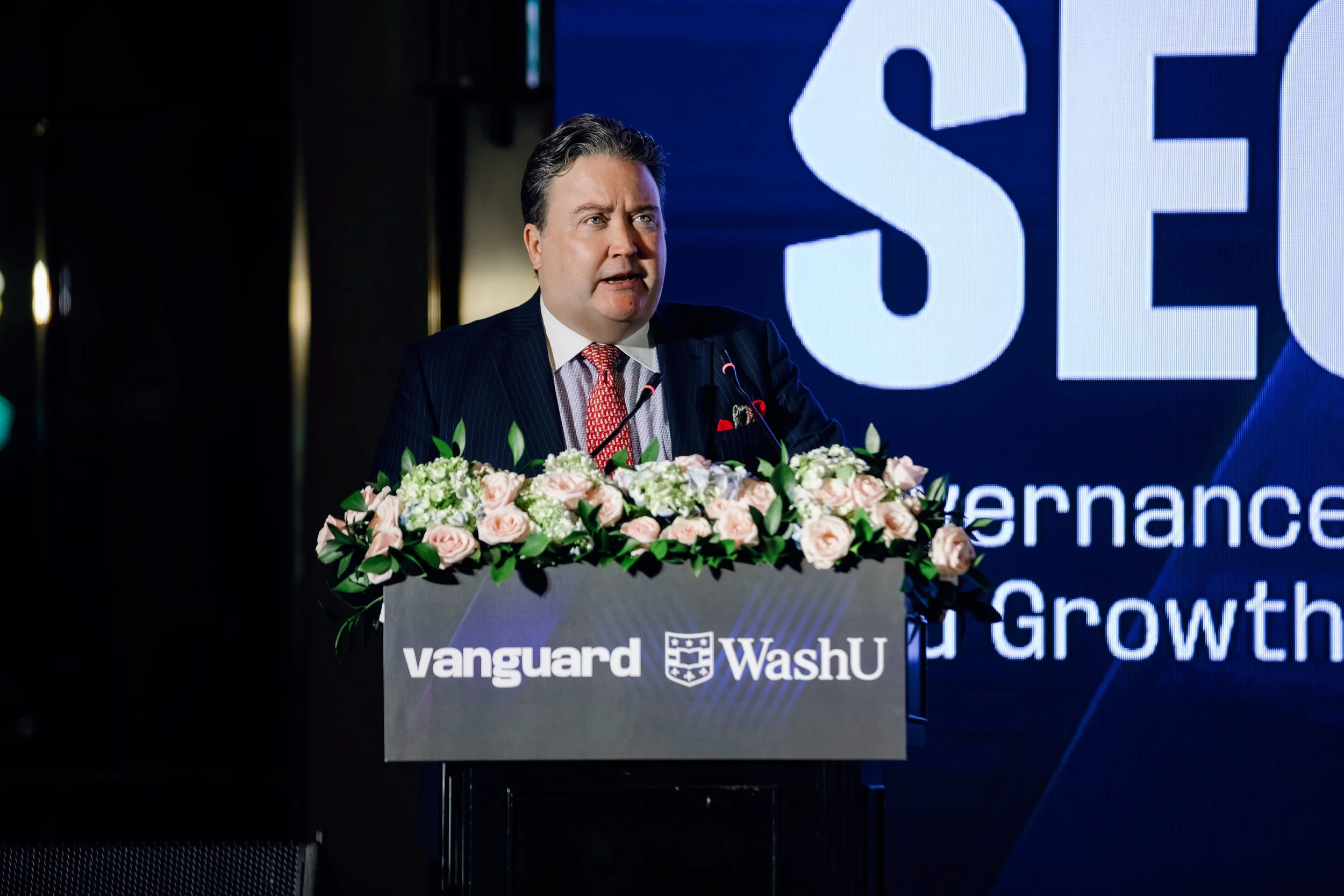A man in a business suit giving a speech at a podium decorated with pink and white flowers. The podium has the logos of Vanguard and WashU. Behind him is a large screen displaying part of the word 'SEC' and some text about governance and growth.