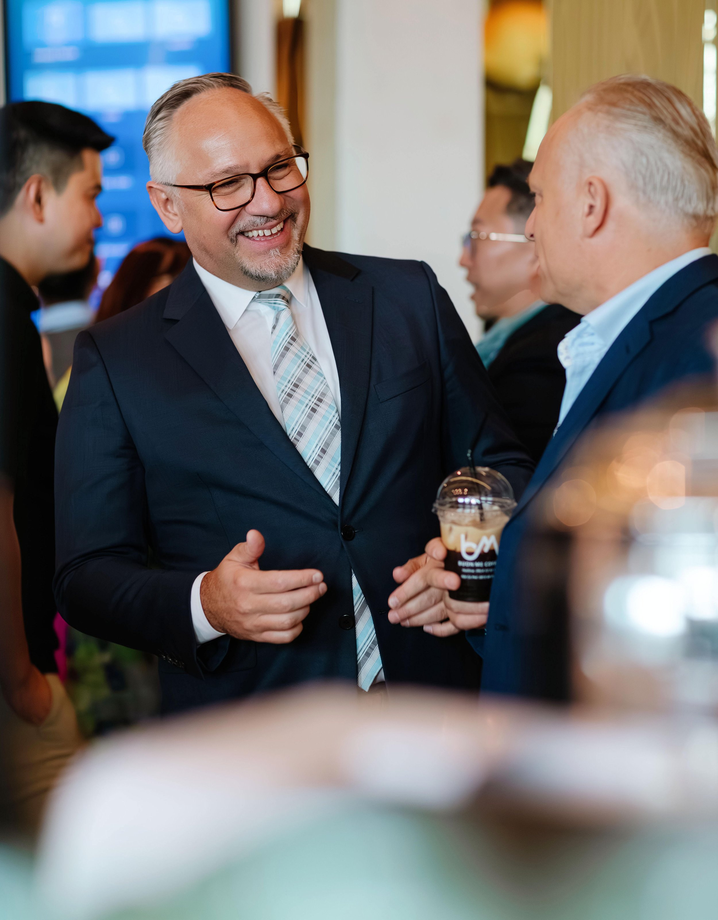 Two men in suits are smiling and talking at a professional event, one is holding a cup of iced coffee.