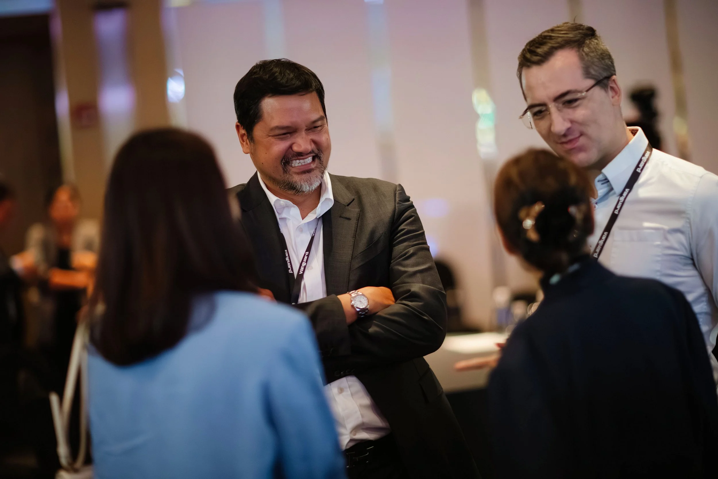 Four professionals, two men and two women, are engaged in a conversation at a conference or seminar. One man is smiling with crossed arms, while the others are listening attentively.