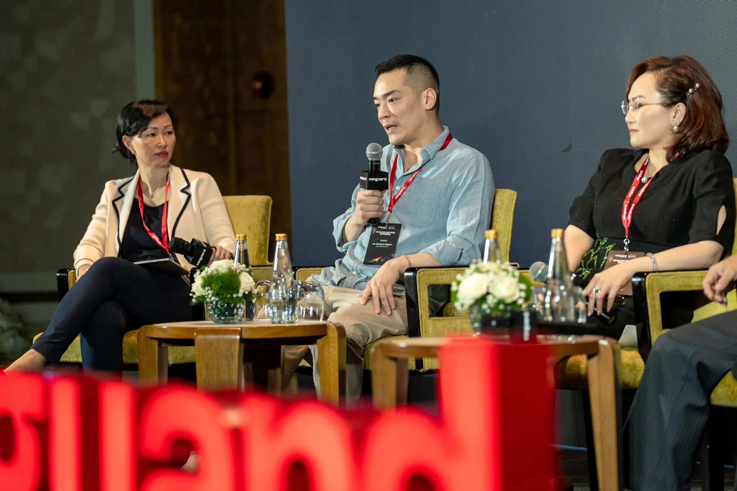 Three people speaking on a panel at a conference, with two women and one man holding a microphone, seated in yellow chairs with small wooden tables in front of them, decorated with vases of white flowers and bottles of water.