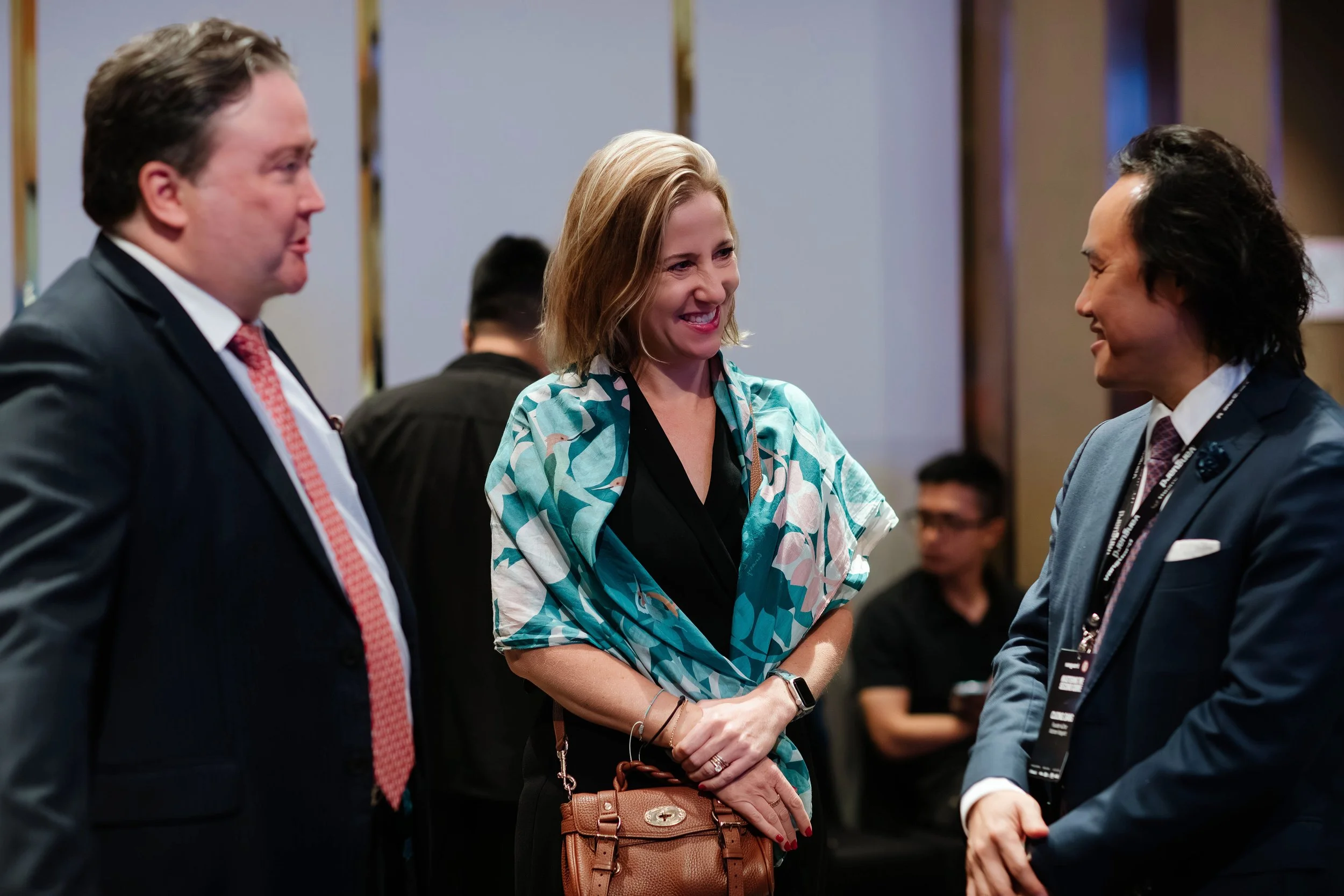 Three people in business attire engaging in conversation at a professional event, two men and a woman, with other attendees in the background.