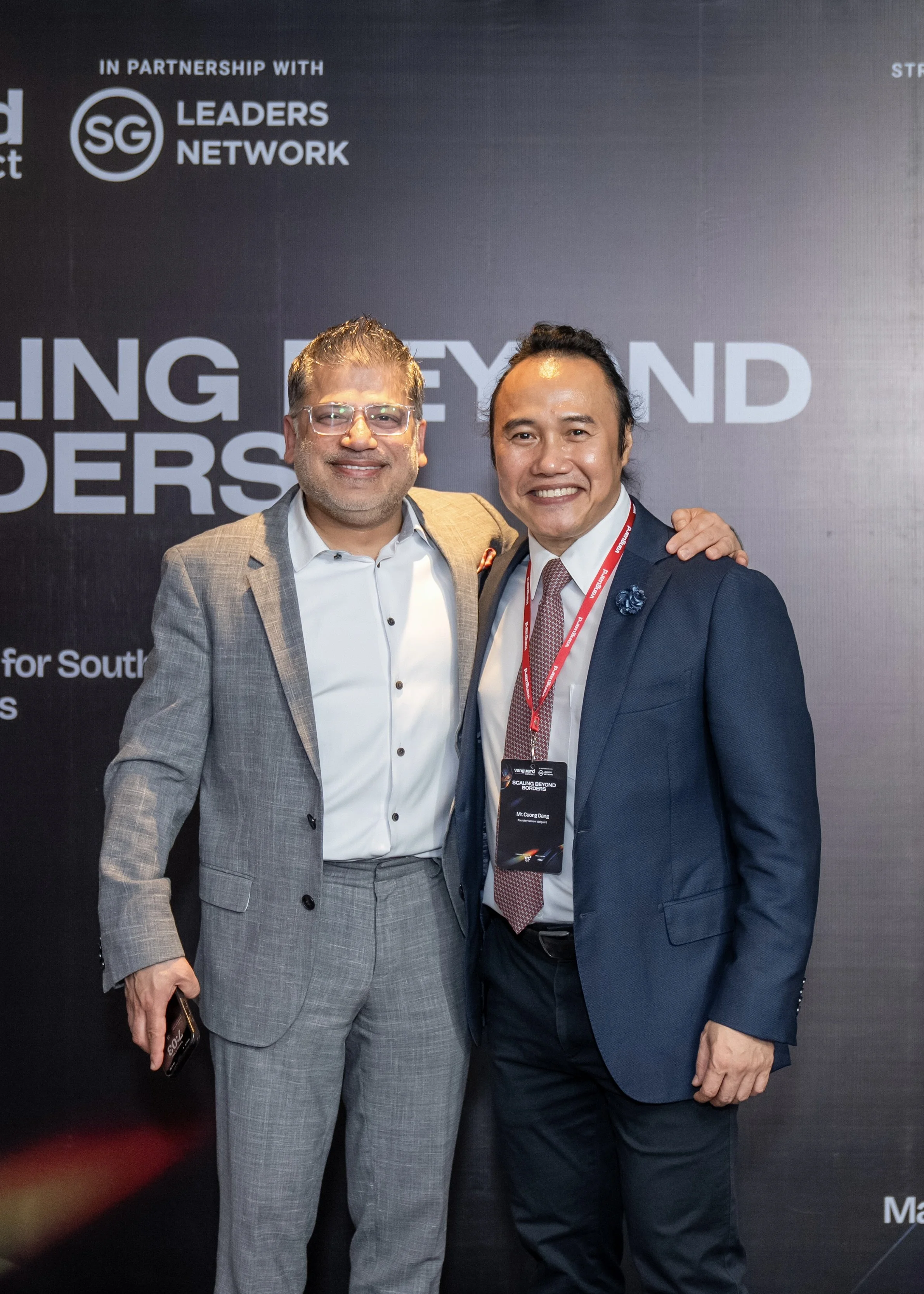 Two men in suits smiling and posing together at a professional event with a black backdrop featuring logos and text.