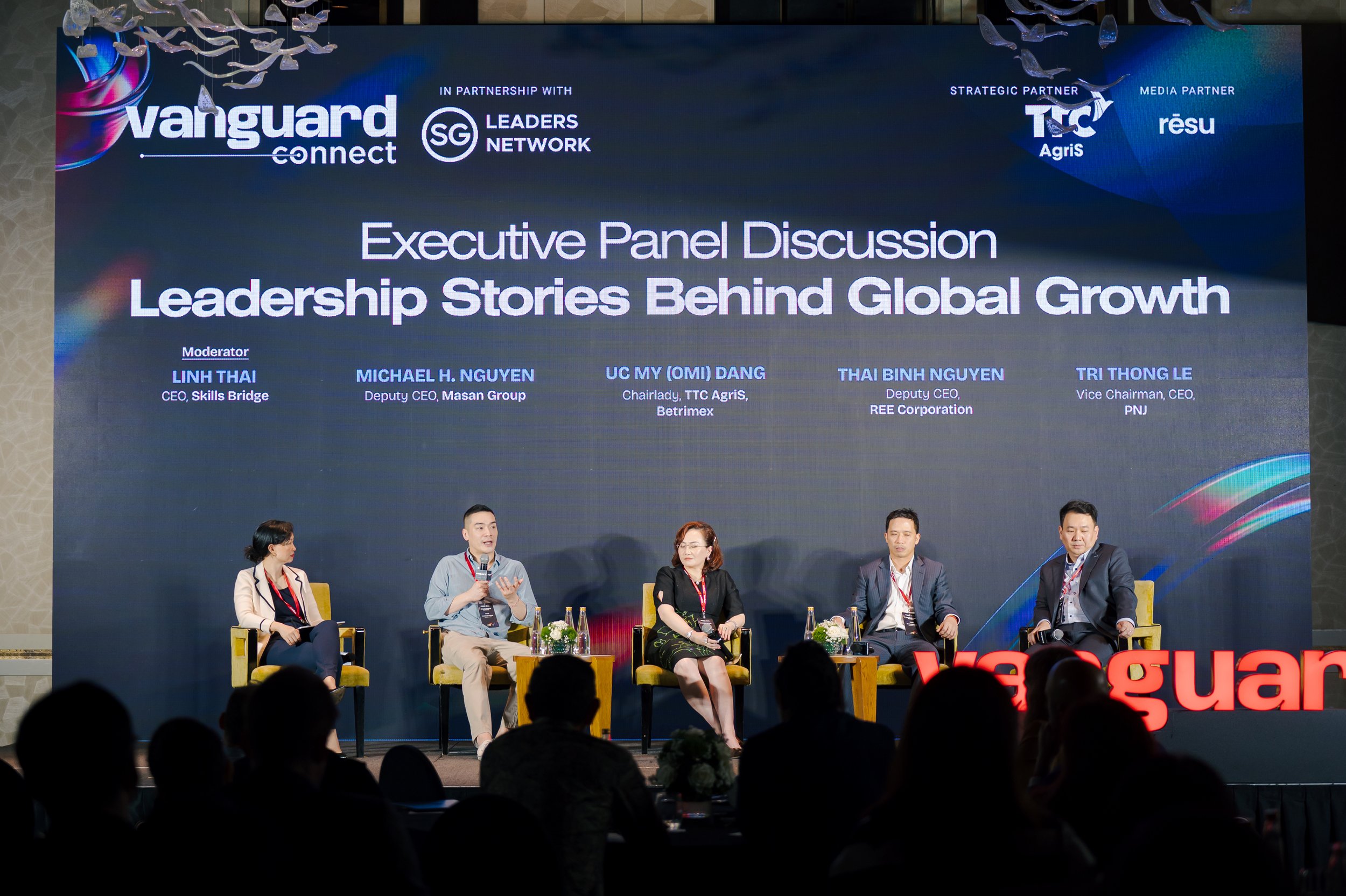 A panel discussion at a conference titled 'Leadership Stories Behind Global Growth' with five speakers seated on stage. The large screen behind them displays the event title, moderator, and panelists' names and titles. Audience silhouettes are visibl