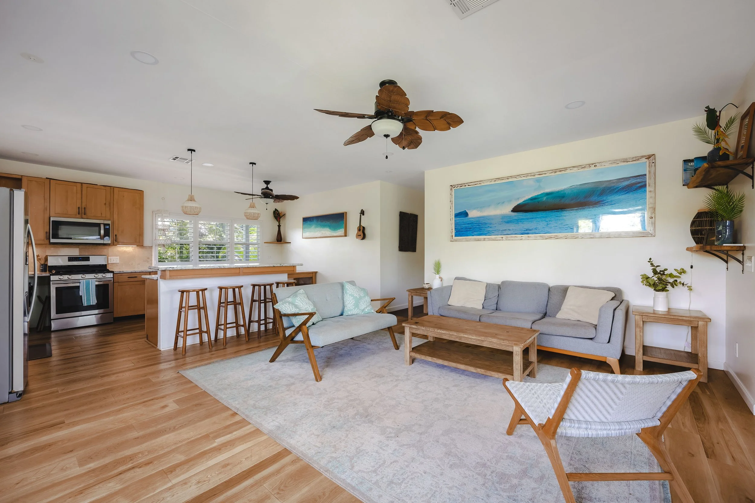 Living room with light-colored walls, a large framed ocean painting, a gray sofa with pillows, a wooden coffee table, and two wooden chairs with woven seats. There are wooden hardwood floors, a ceiling fan with leaf-shaped blades, and open kitchen wi