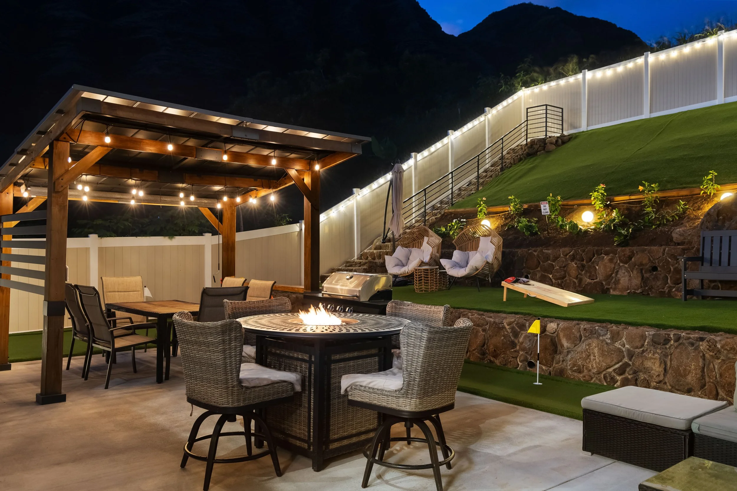 Nighttime outdoor backyard patio area with a fire pit, tables, chairs, string lights, and a fenced grassy slope with steps leading up.