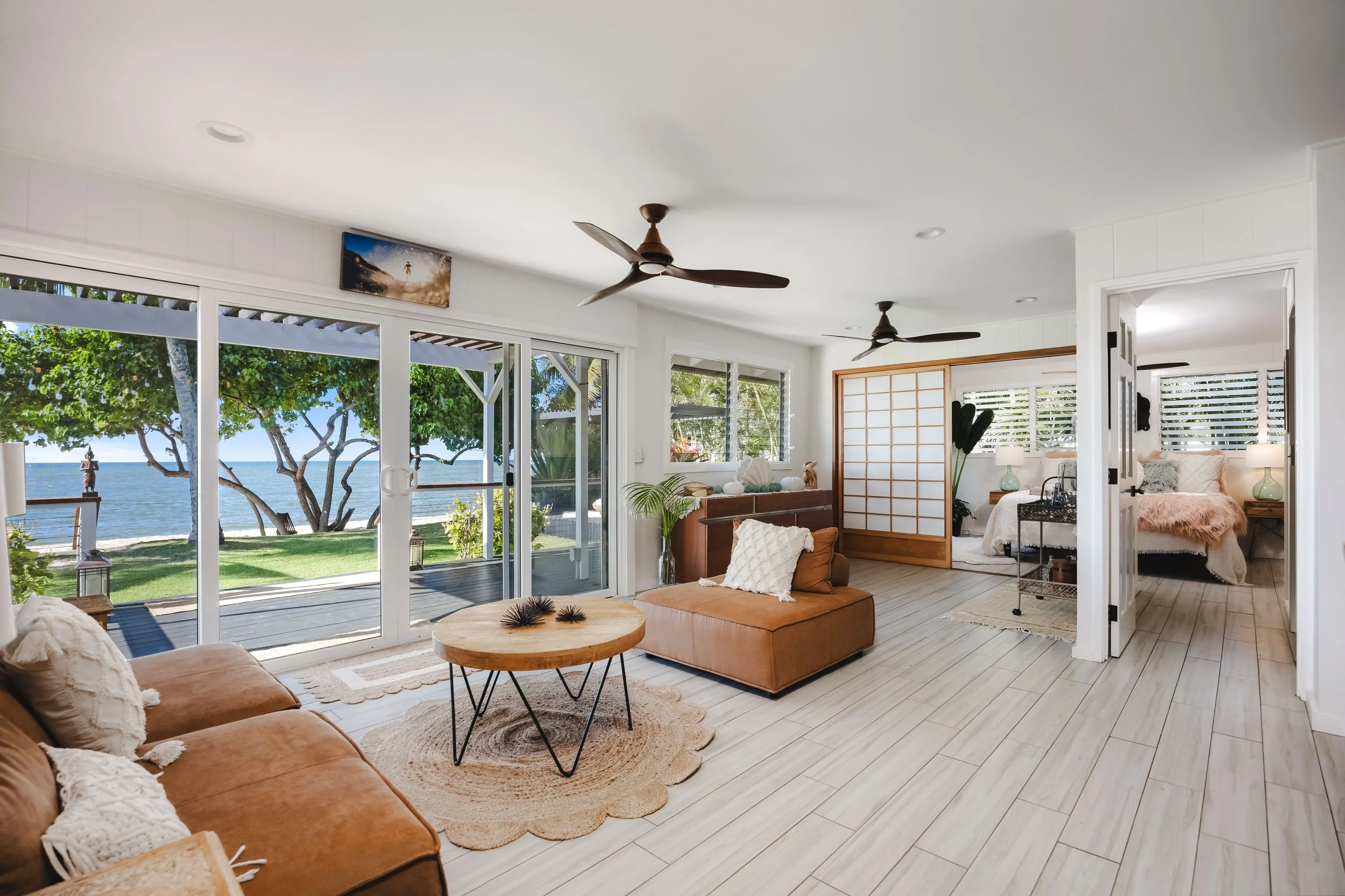 Bright living room with large sliding glass doors showing ocean view, white walls, ceiling fans, beige sofa, round wooden coffee table, plants, and a bedroom partially visible through an open doorway.