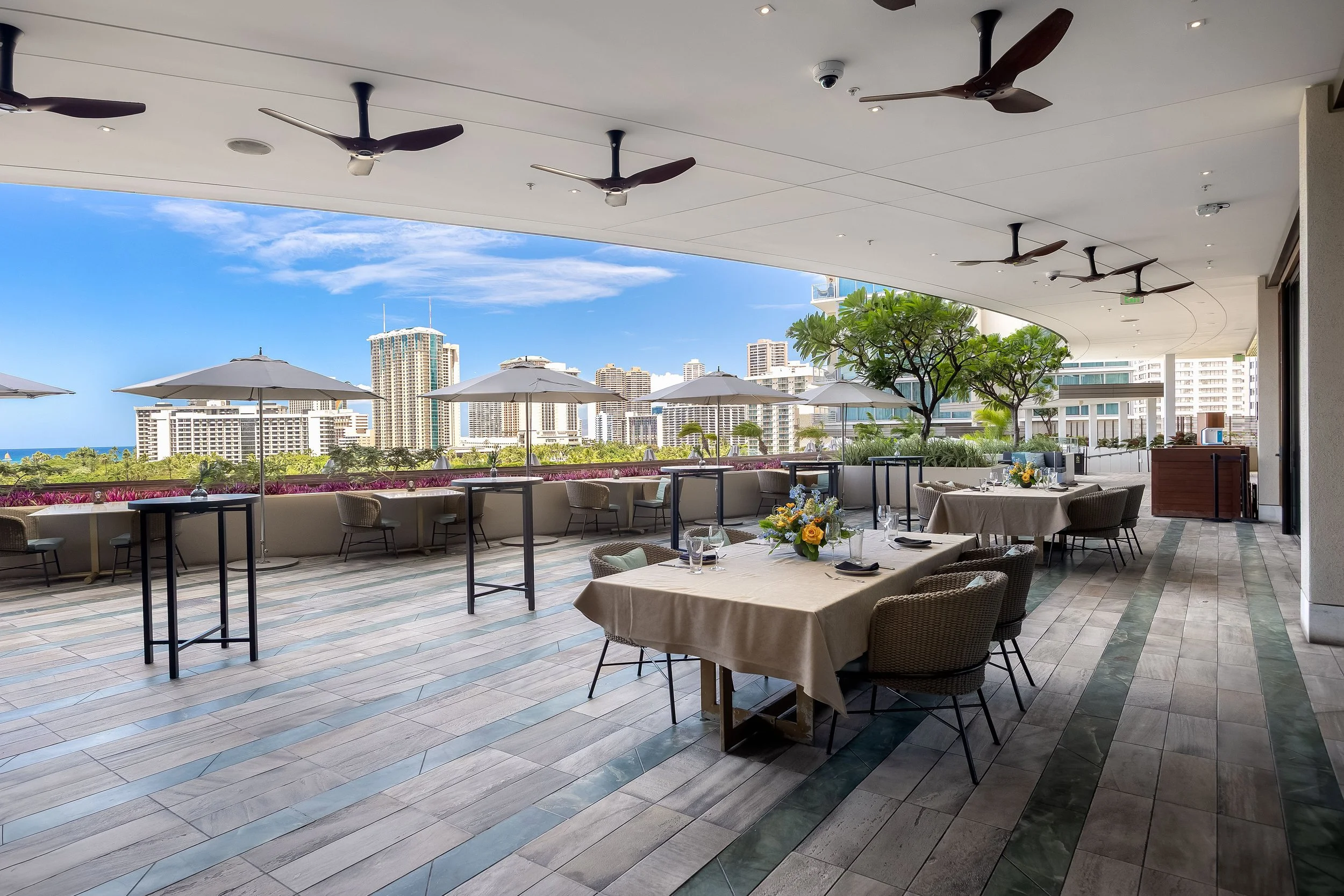 An outdoor dining area on a terrace with tables and chairs, decorated with flowers, and overlooking a city skyline with high-rise buildings and a blue sky.