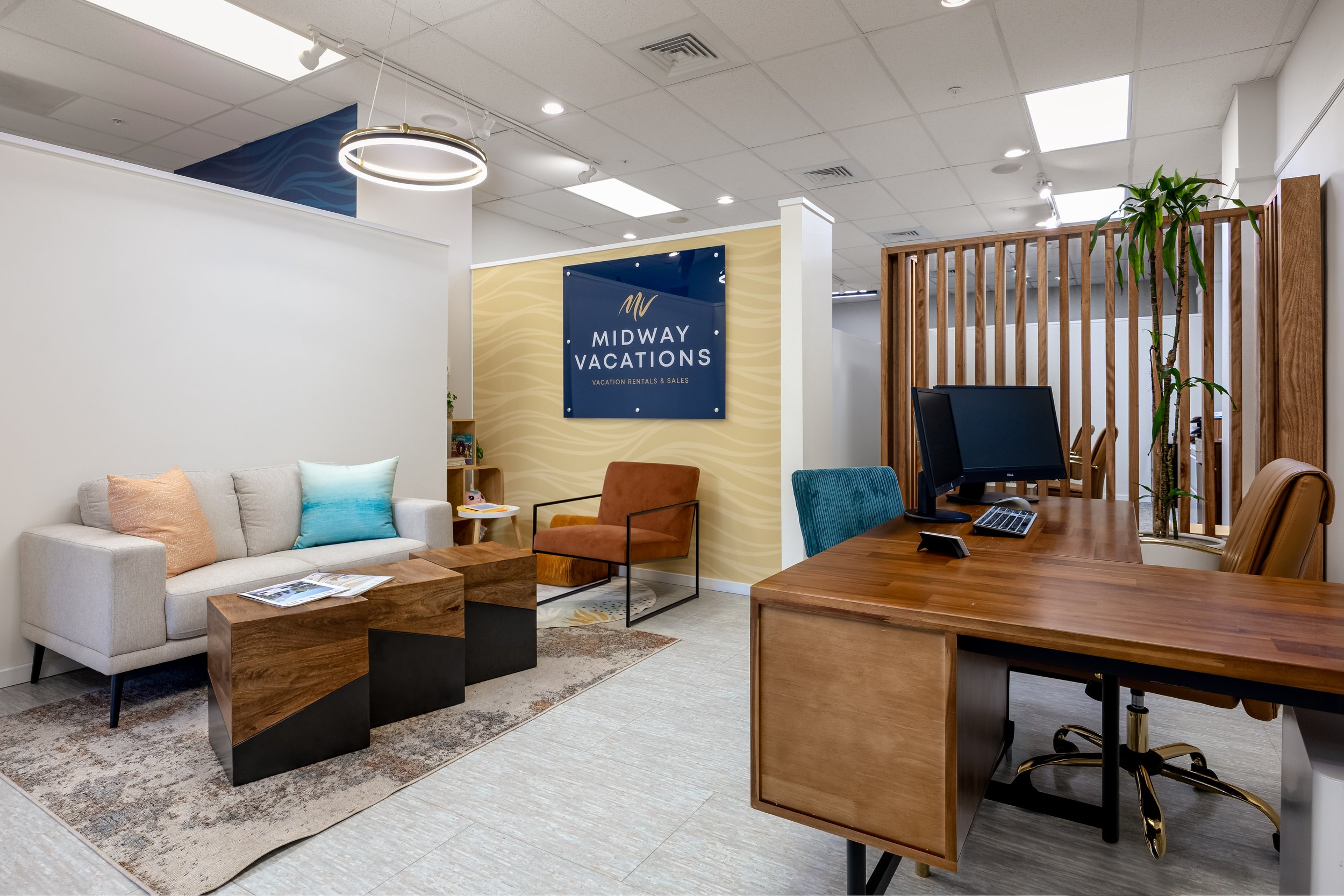 Office reception area with a white sofa, colorful cushions, wooden tables, a computer station, and a sign for Midway Vacations, featuring modern furniture and decor.