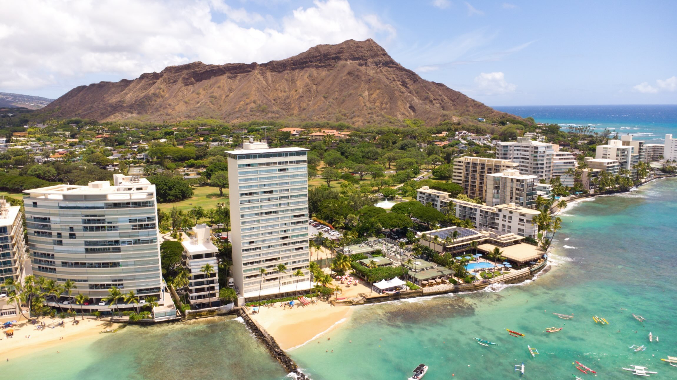 Aerial view of a cityscape with modern high-rise buildings beside a beach and ocean, a lush green mountain in the background, partly cloudy sky.