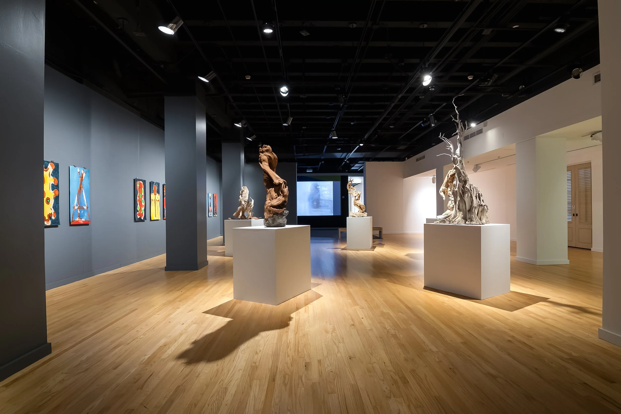 An art gallery with colorful paintings on the gray wall and multiple sculptures on white pedestals, illuminated by spotlights in the ceiling, and light wooden flooring.