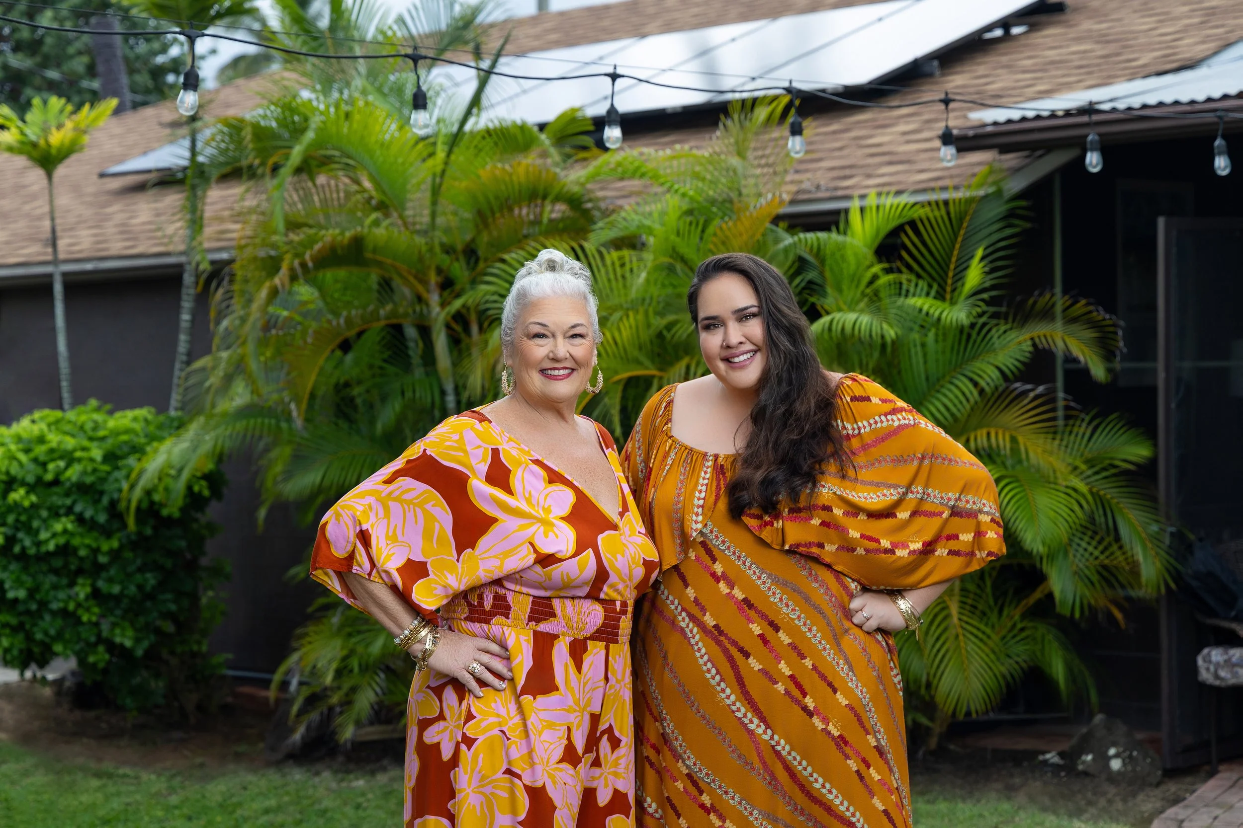 Two women standing outdoors in front of tropical plants and a house, smiling at the camera. Both are wearing colorful patterned dresses and jewelry.