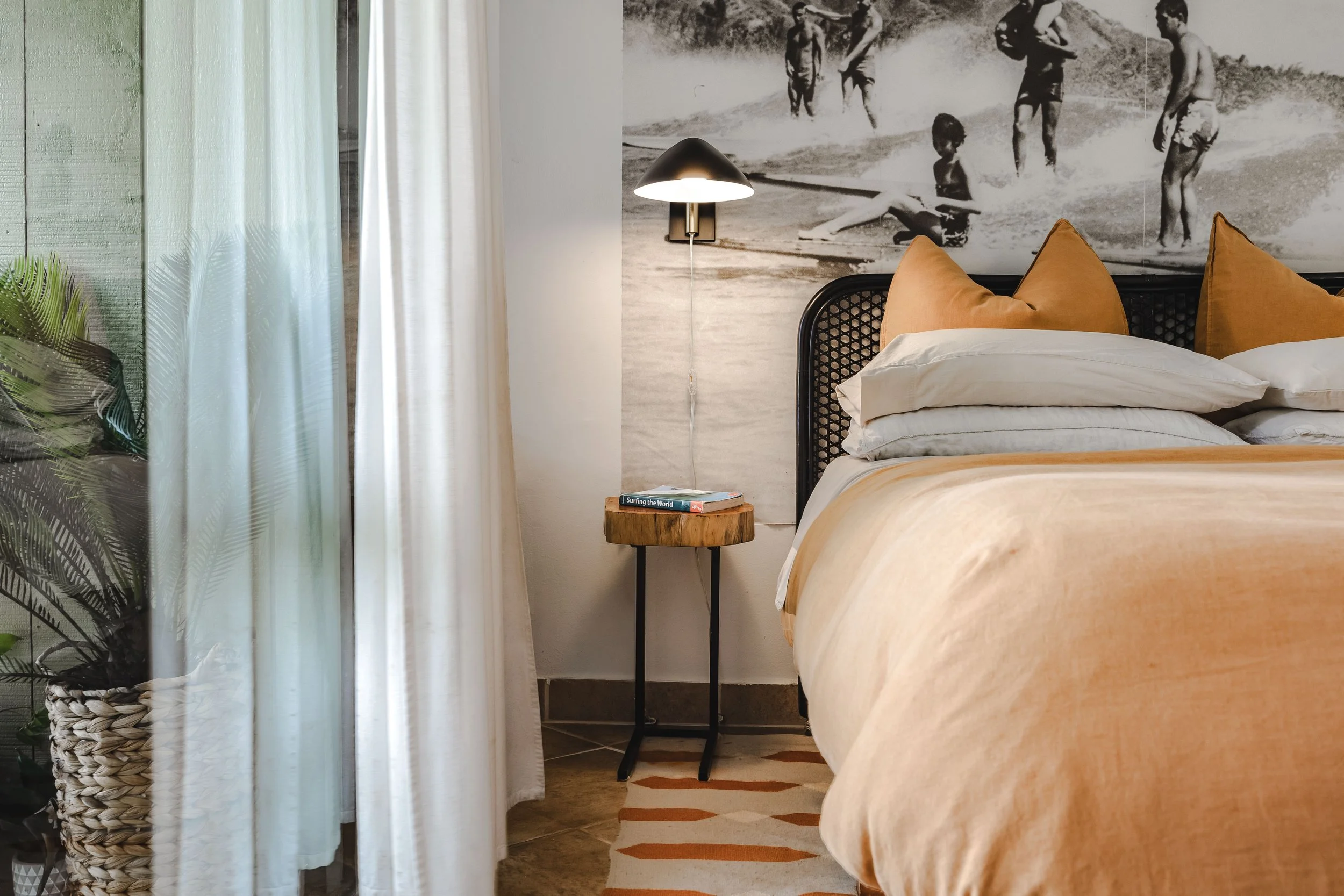 A bedroom with an orange bed, white pillows, a wooden side table with books, a black and white beach-themed mural, a wall lamp, white curtains, and a plant in a woven basket.