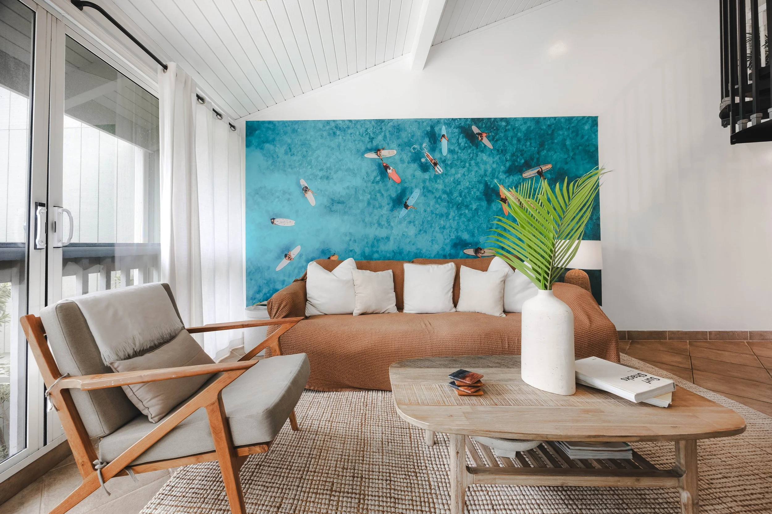 Living room with white walls and ceiling, featuring a large mural of people surfing in blue water on the main wall. There is a brown sofa with white cushions, a white concrete vase with green palm leaves, a wooden coffee table with magazines, and nea