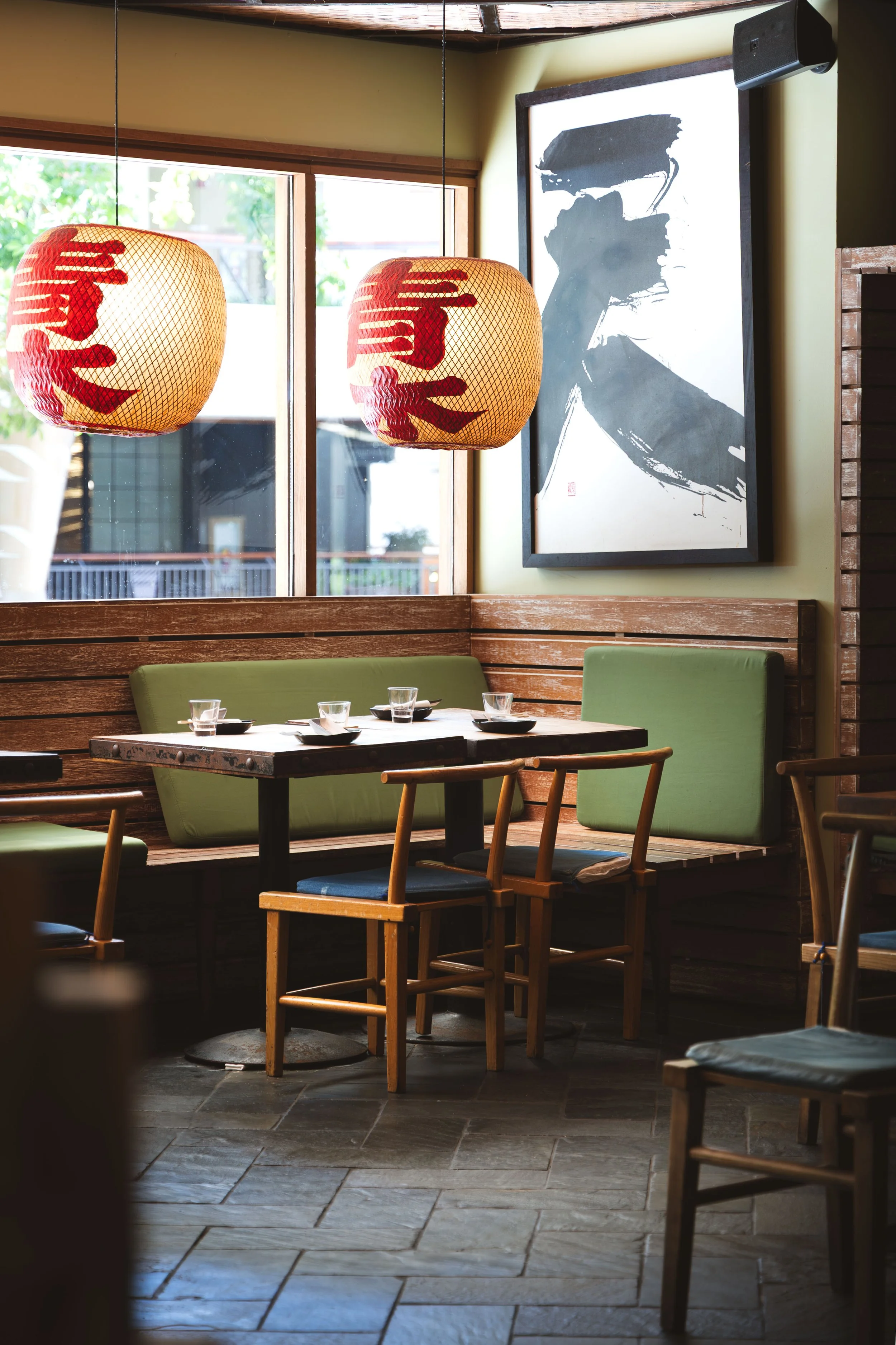 Interior of a restaurant with wooden furniture, green cushioned chairs, and hanging paper lanterns with Japanese characters. A large black and white portrait hangs on the wall near a window, with sunlight streaming in.