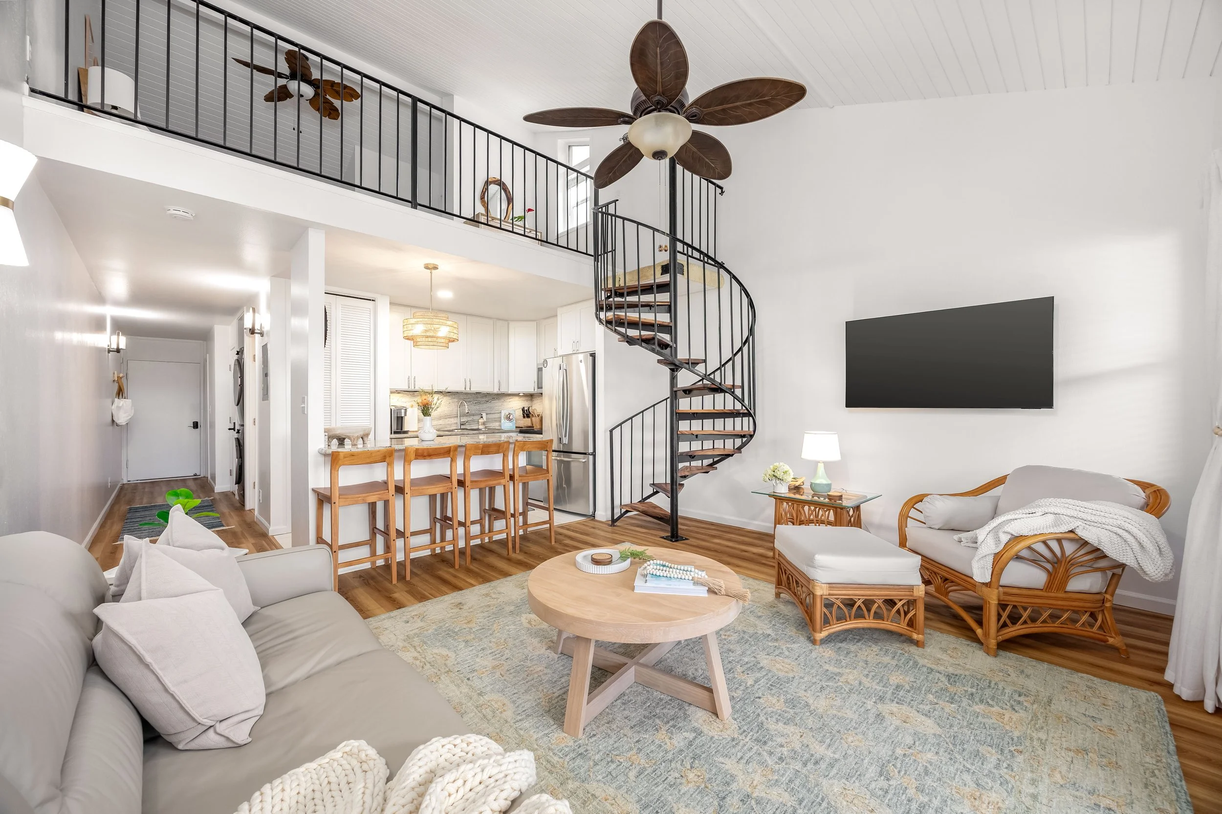 Living room with white walls, beige sofa with white pillows, rattan chairs, a round coffee table, a television on the wall, a staircase with black railing, and a loft area with ceiling fan, decorated with flowers and art.