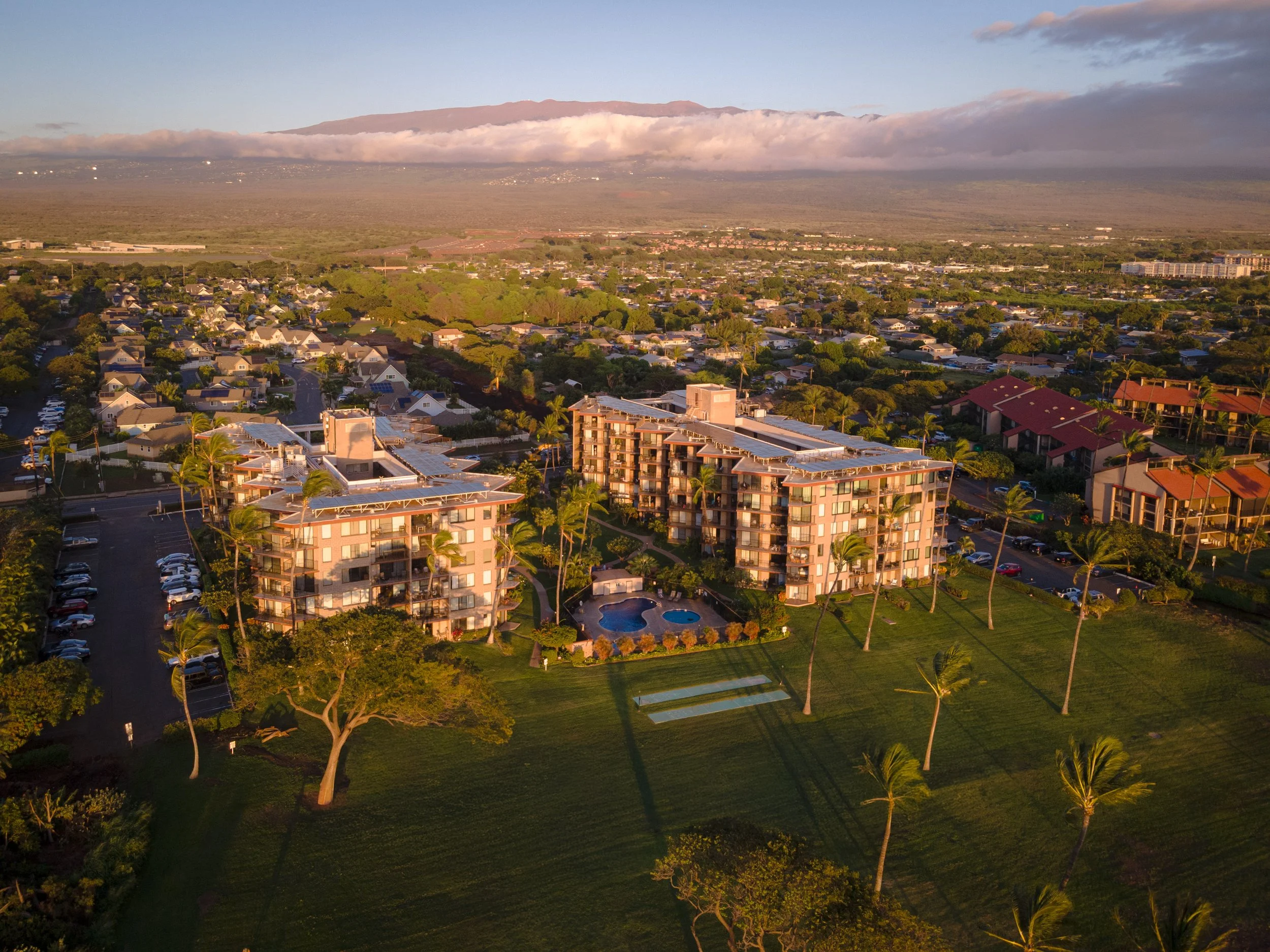 Aerial view of a residential area with multi-story apartment buildings, surrounded by trees and a golf course, with a mountain in the background under a partly cloudy sky.