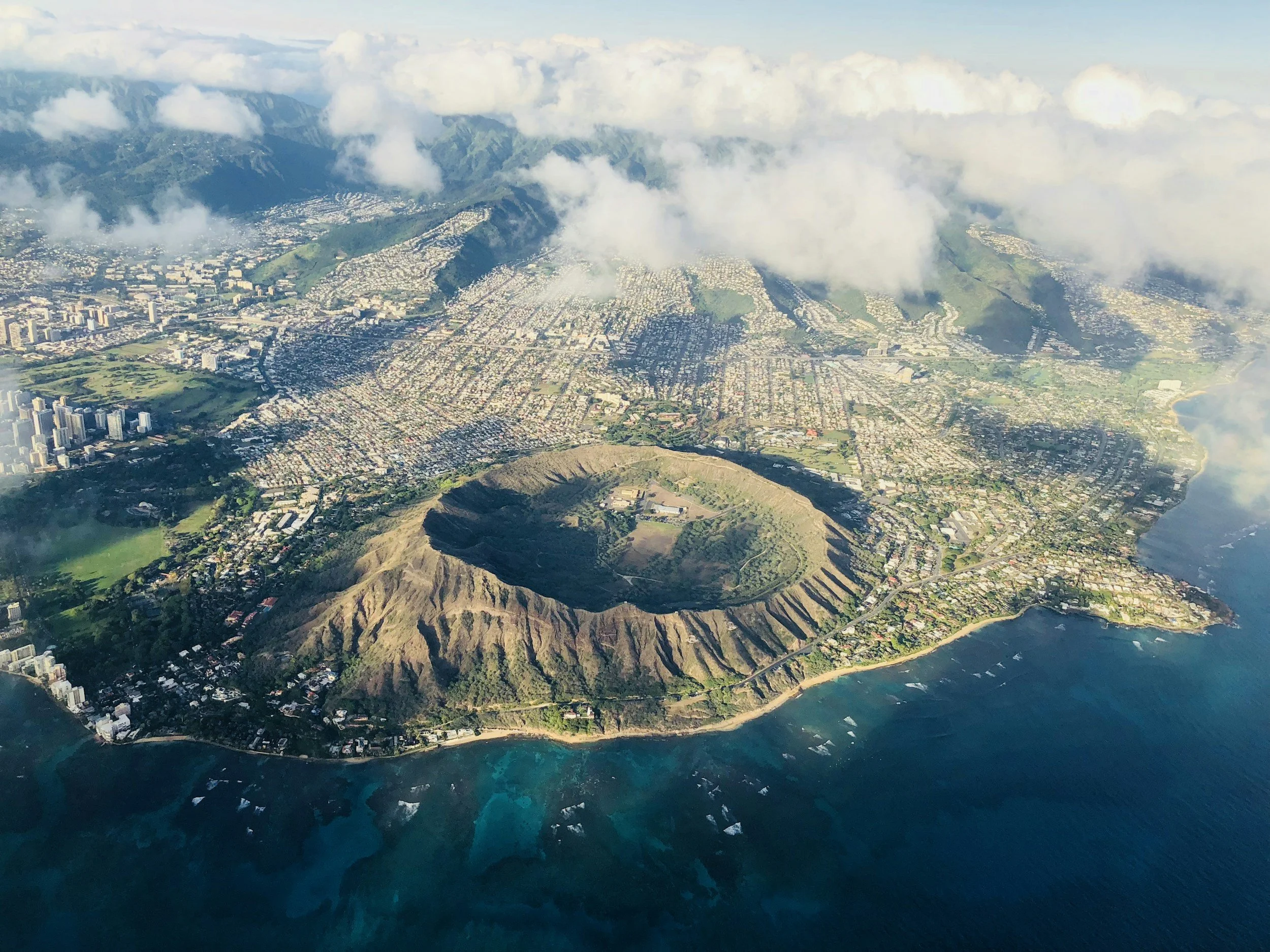 An aerial view of a coastal city with mountains in the background, partially obscured by clouds.
