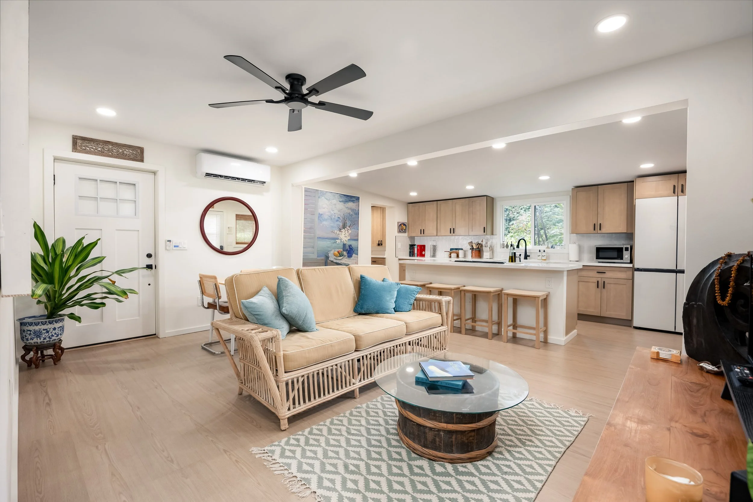 Open-concept living room and kitchen with beige sofa, wicker furniture, green plant, wall art, and kitchen appliances.