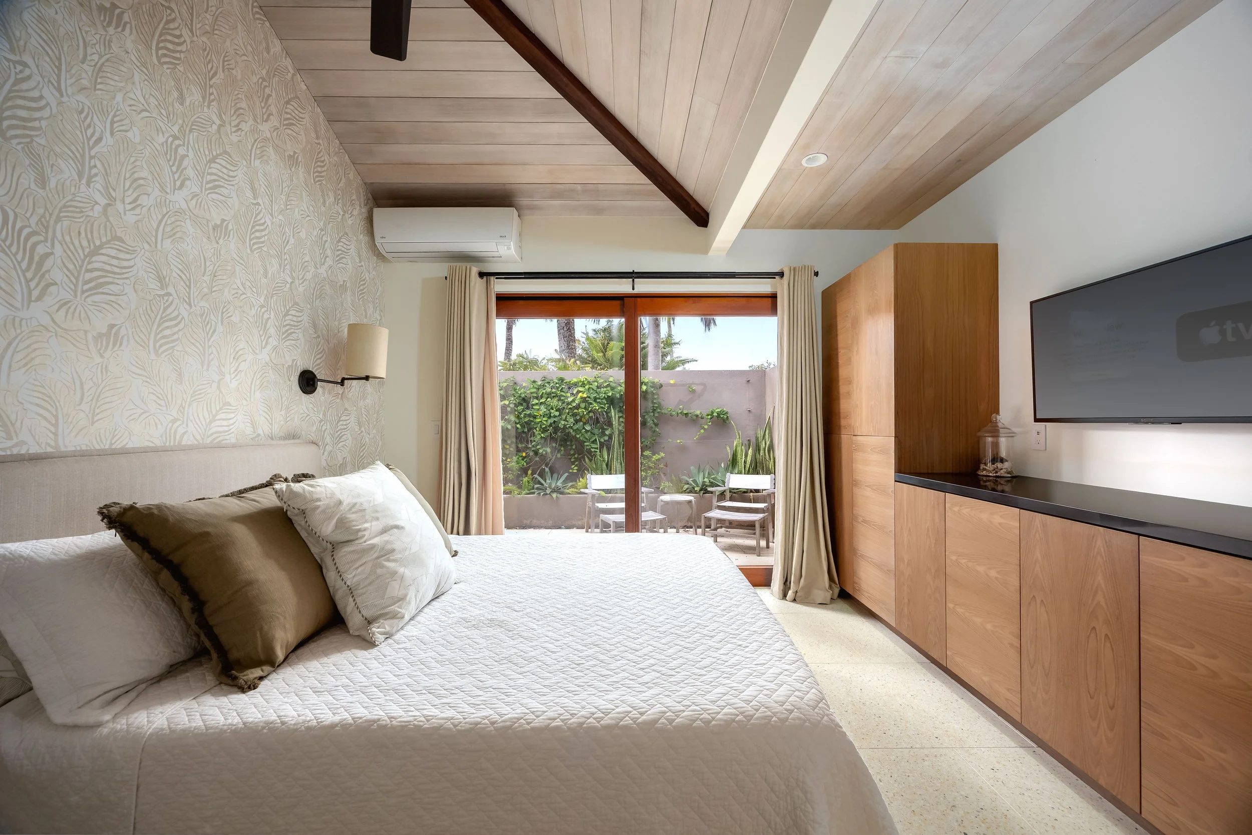 A bedroom with a bed, pillows, wall-mounted light, window with curtains, and patio with chairs and greenery outside.