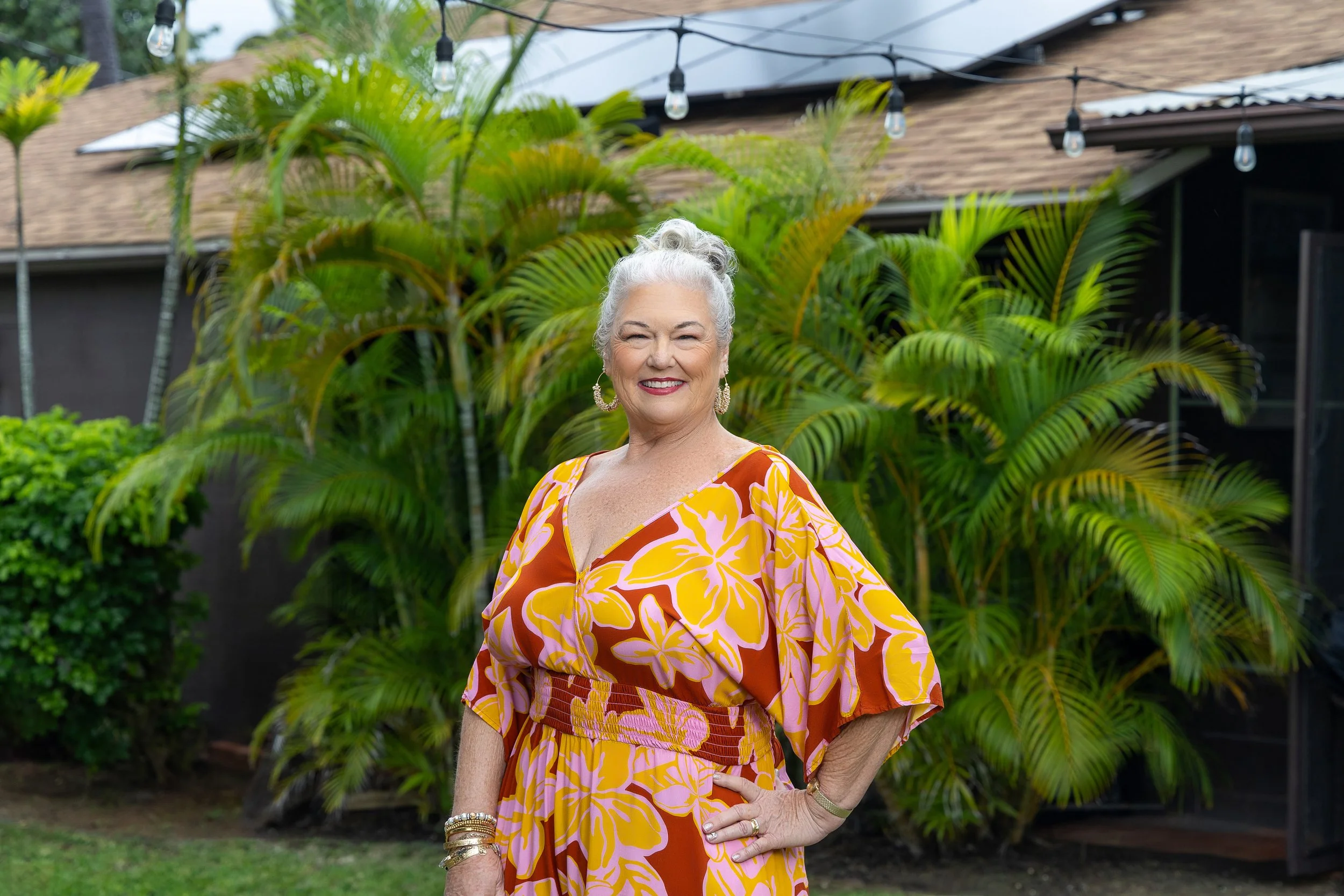 An older woman with white hair styled in a bun, smiling, wearing a bright orange and yellow floral dress, standing outdoors in front of green tropical plants and a house with string lights overhead.