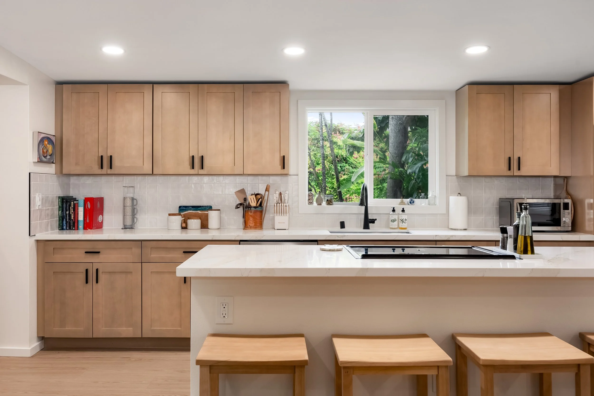 A modern kitchen with beige wooden cabinets, a white countertop, and a large window showing green trees outside. The kitchen has various utensils, books, and decorative items near the window, with a built-in stove on the island and three wooden stool