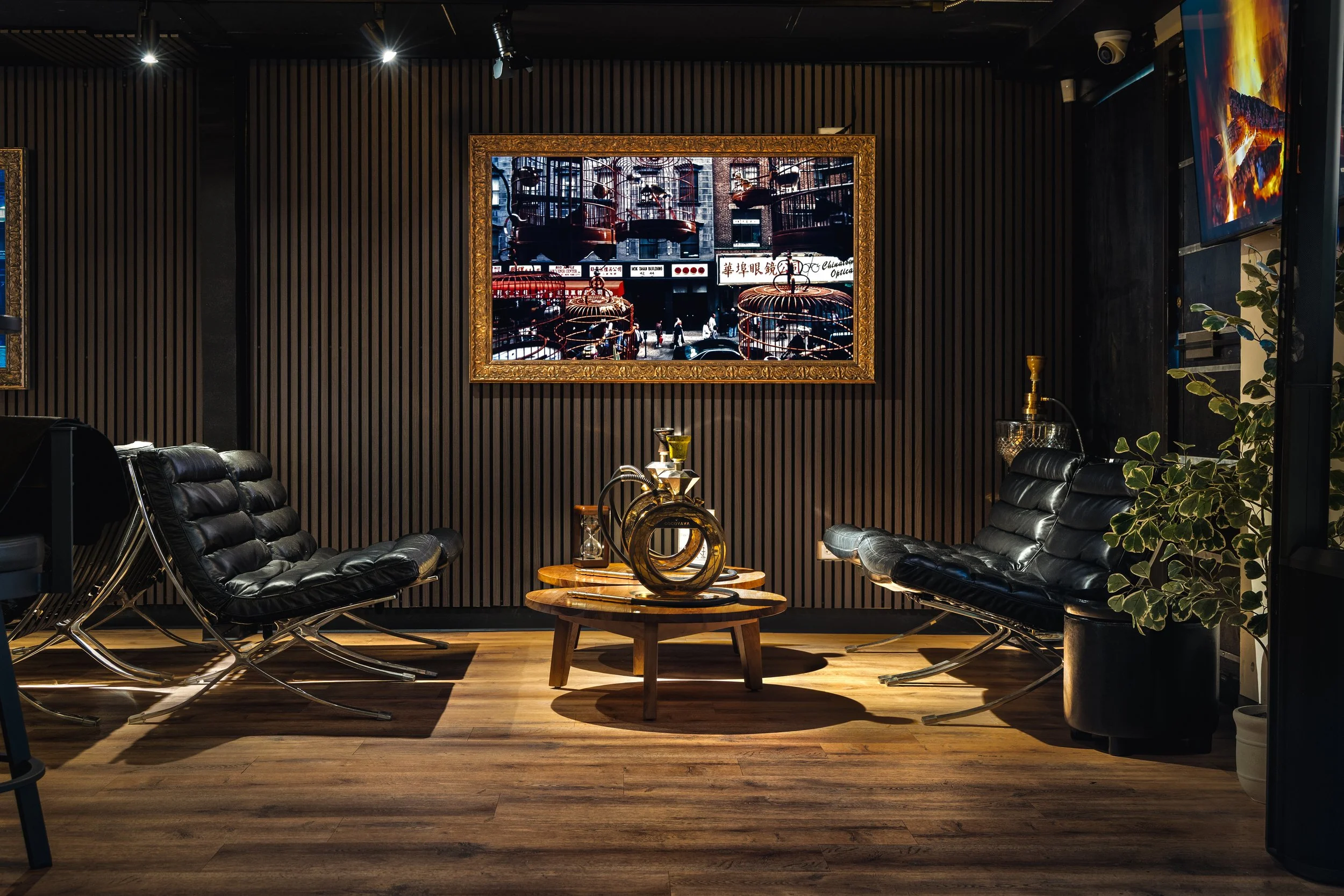 A modern lounge with two black leather chairs, a small wooden table with decorative objects, a large framed city scene photograph on the dark wood-paneled wall, and green foliage to the right.