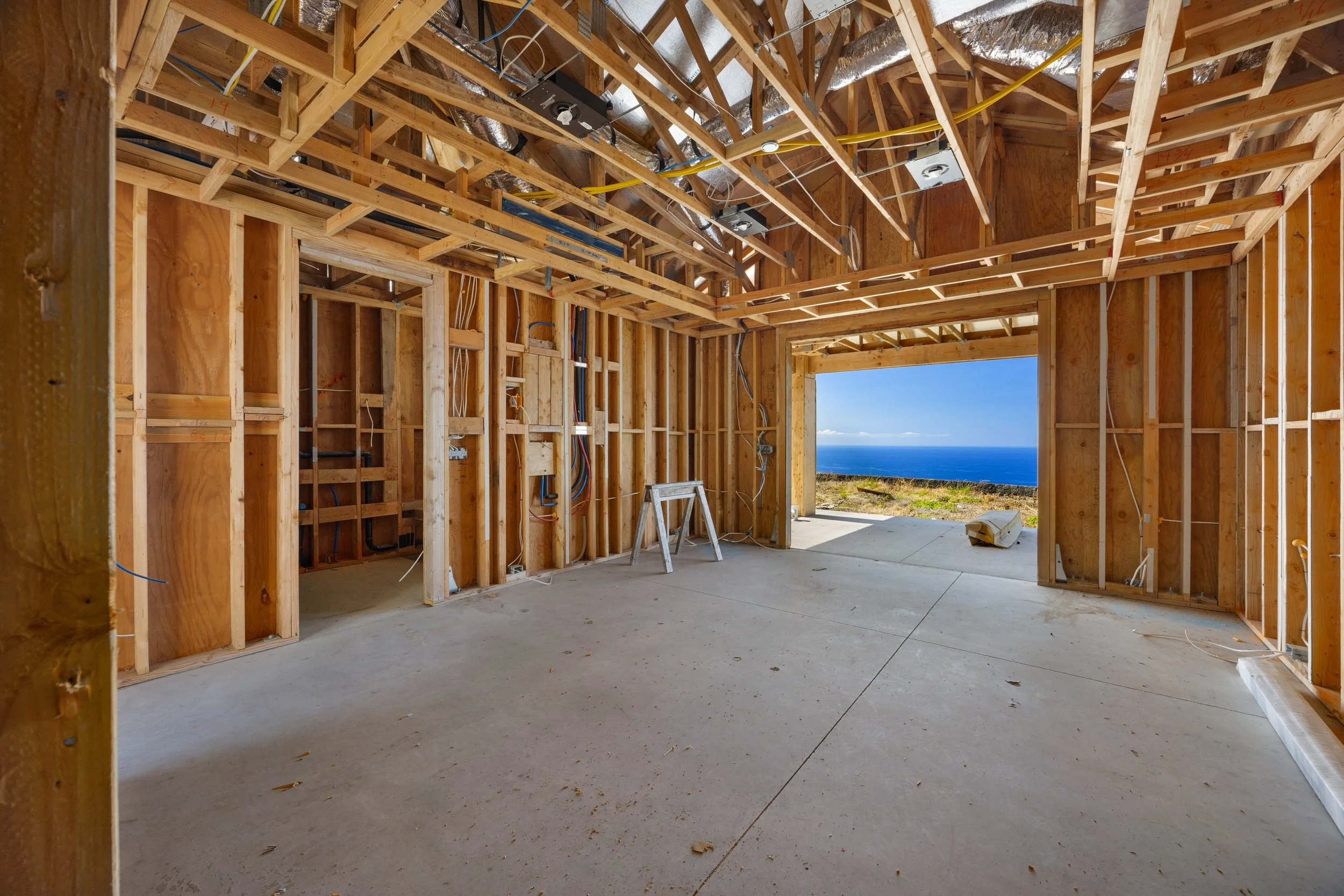Interior view of a house under construction with exposed wooden framing, electrical wiring, HVAC ducts, and a large opening showing ocean view.