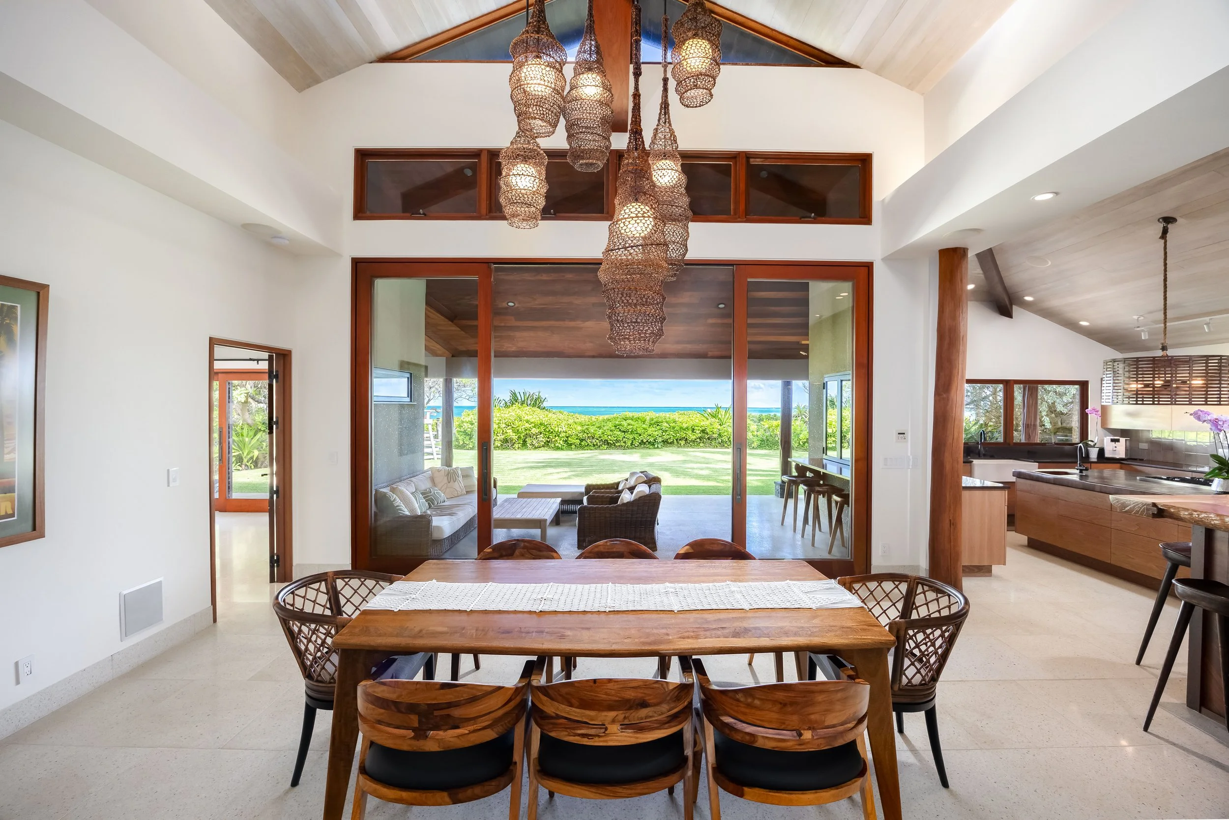 Open living and dining area with view of outdoor greenery and ocean, featuring wooden accents, wicker chairs, and a modern chandelier.