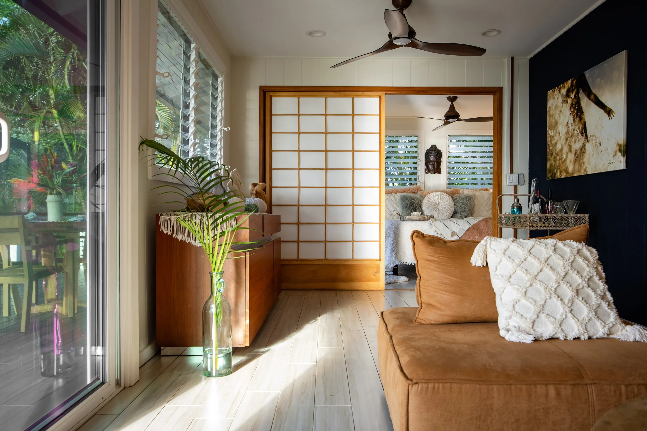 Living room with glass sliding door, wooden floor, plants, and seating area with cushions.