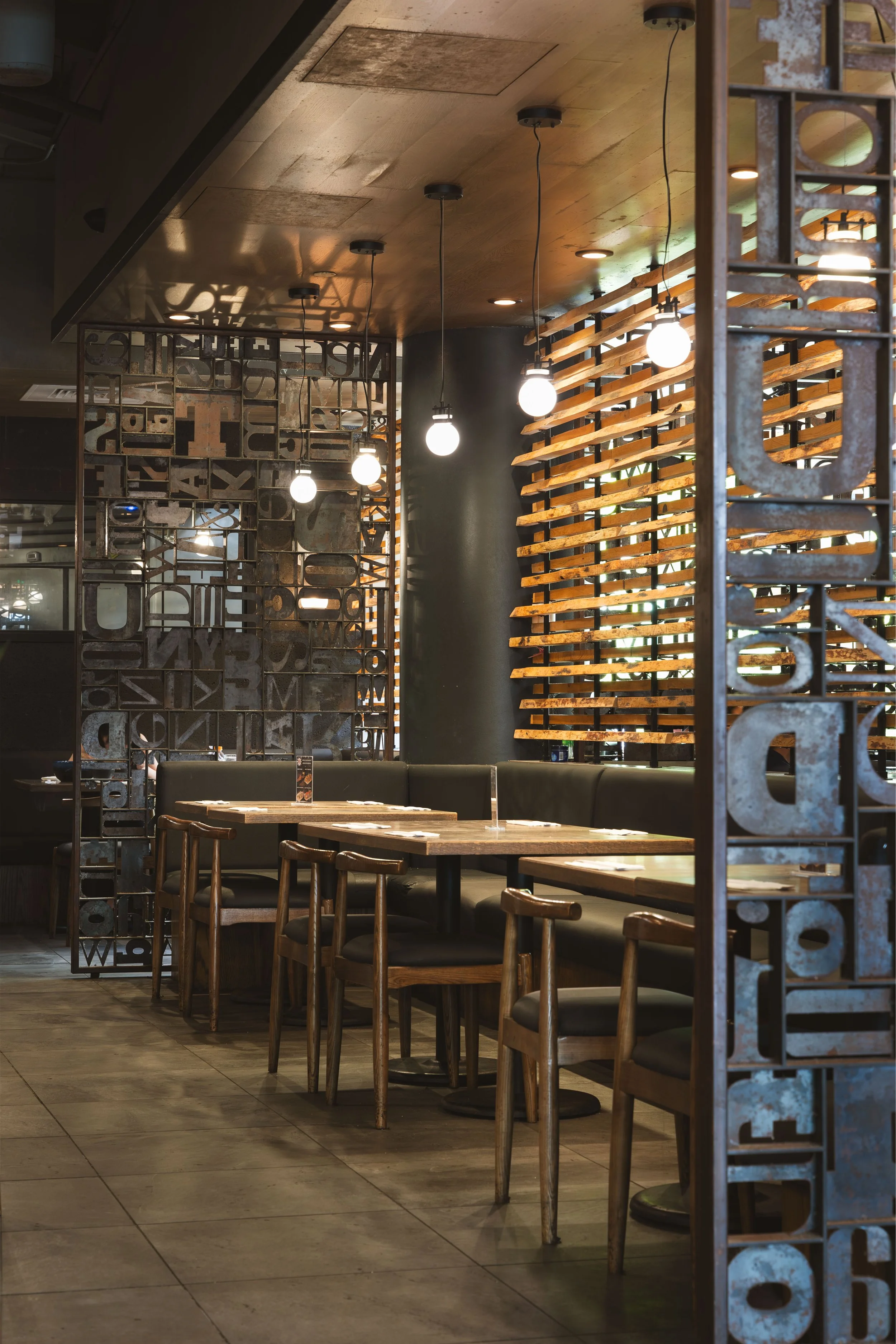 Interior of a modern restaurant with wooden tables, chairs, and decorative metal partition with letters and numbers. Hanging pendant lights illuminate the space, which features a mix of wood and metal design elements.