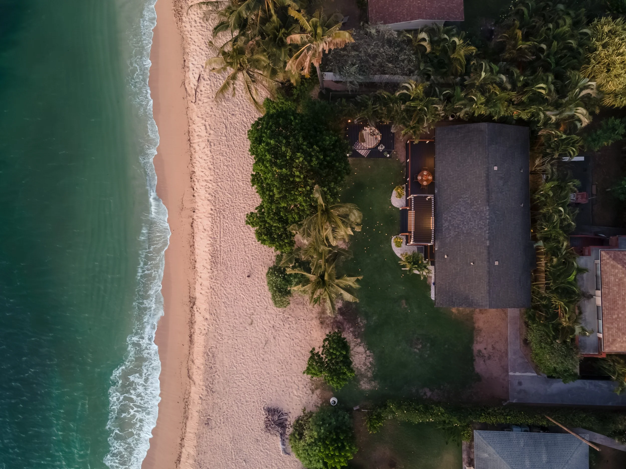 An aerial view of a beachside property with sandy shores, palm trees, a deck with outdoor furniture, and a house surrounded by greenery, taken at dusk.